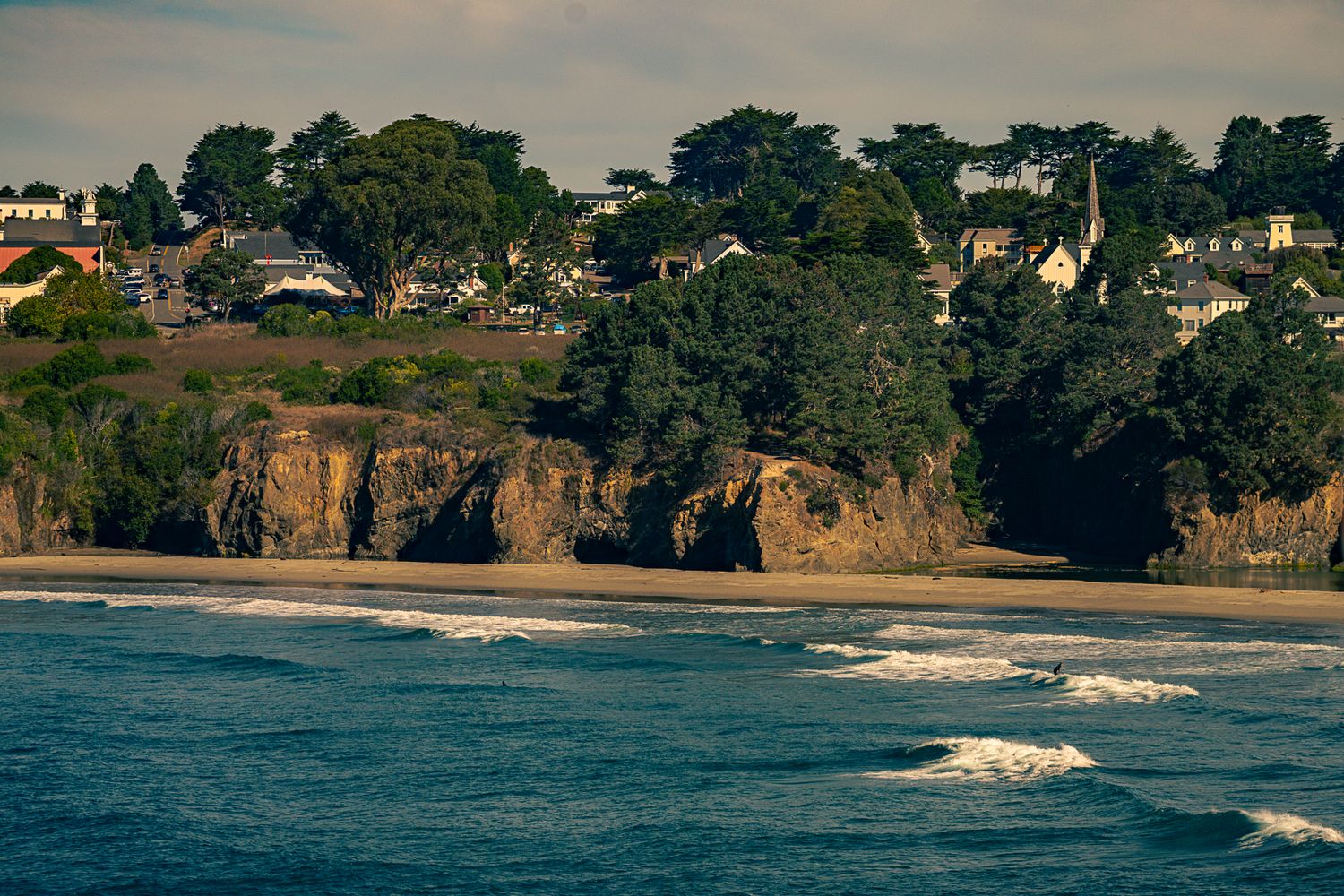 Coastal cliffs with sandy beach, ocean waves, and tree-covered bluff with houses in the background.