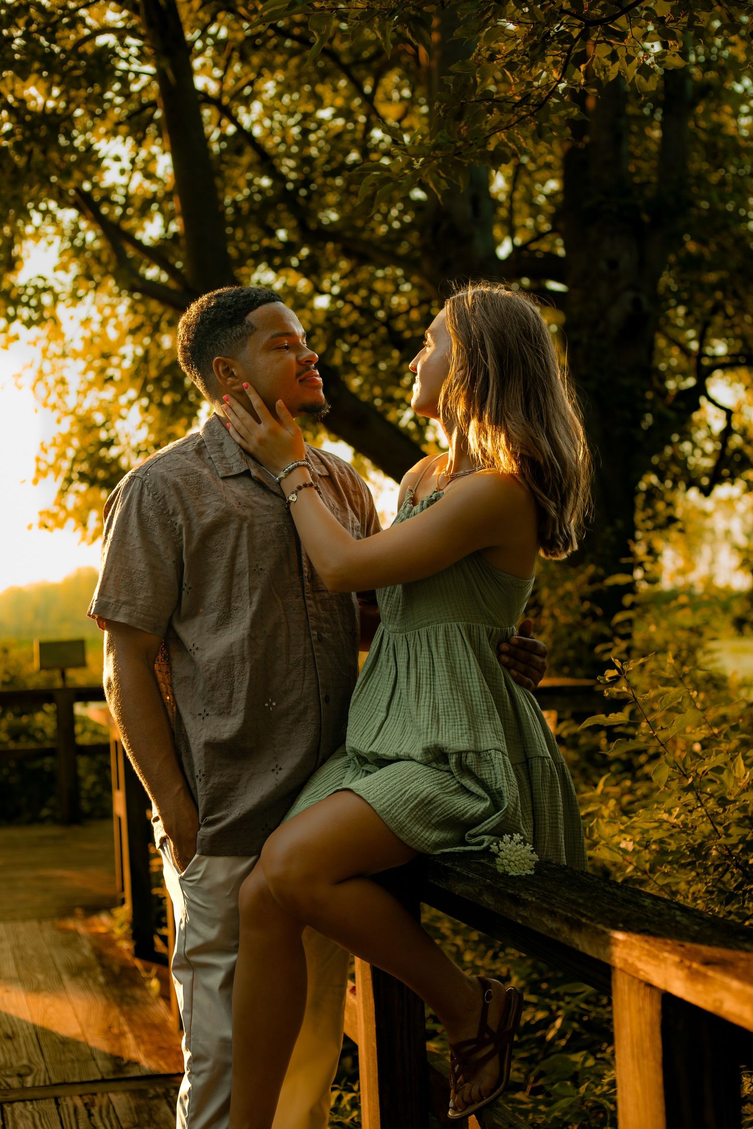 Couple sharing a romantic moment at golden hour on a wooden deck surrounded by lush green trees.