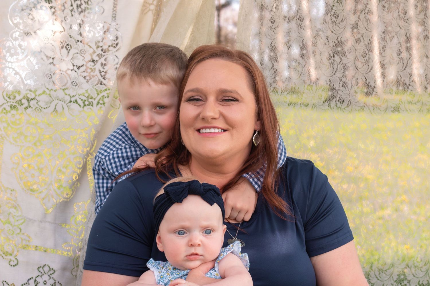A smiling mother holds a baby girl in a blue outfit while a young boy hugs her from behind.