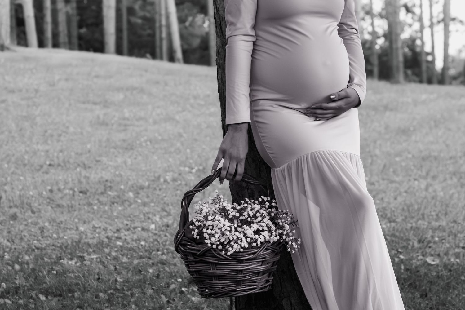 Black and white photo of a pregnant woman in a flowing dress holding a wicker basket of flowers outdoors.