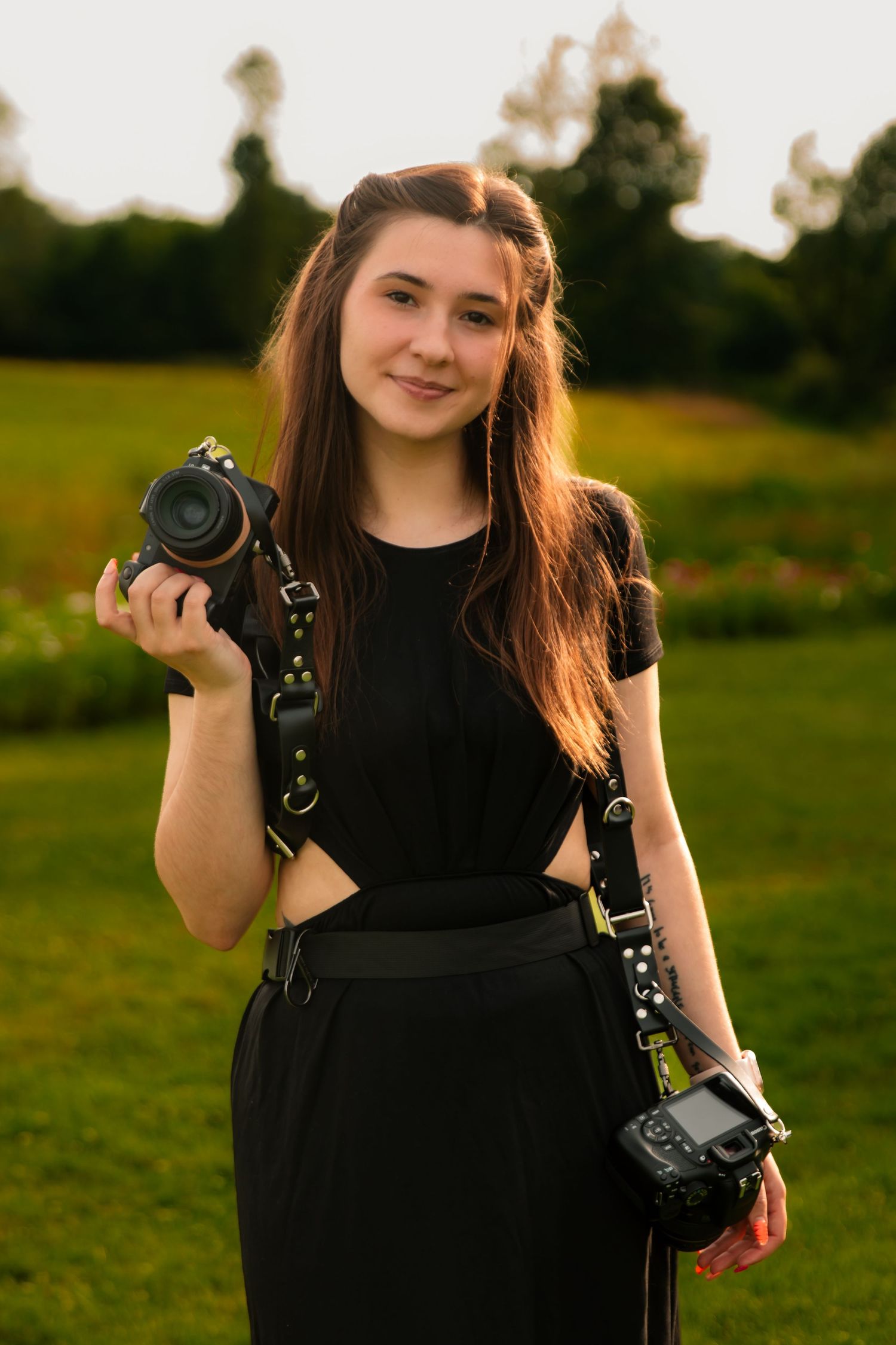 Young woman photographer in black outfit holding two DSLR cameras outdoors in a green park at golden hour.