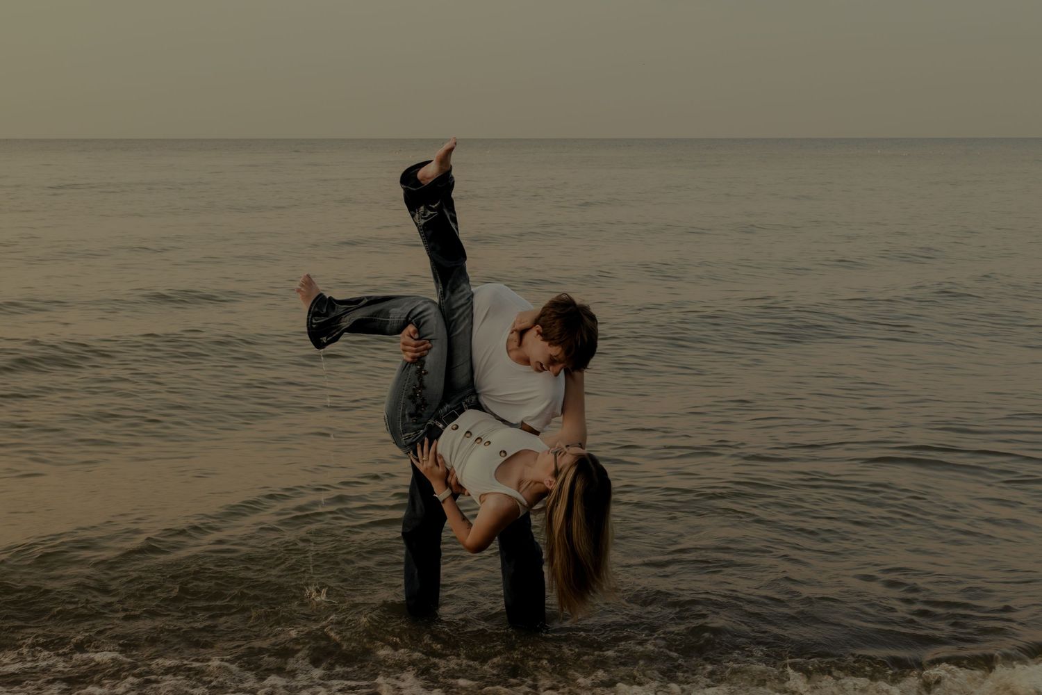 Couple sharing a romantic dip dance pose on a misty beach at dusk, waves gently lapping around them.