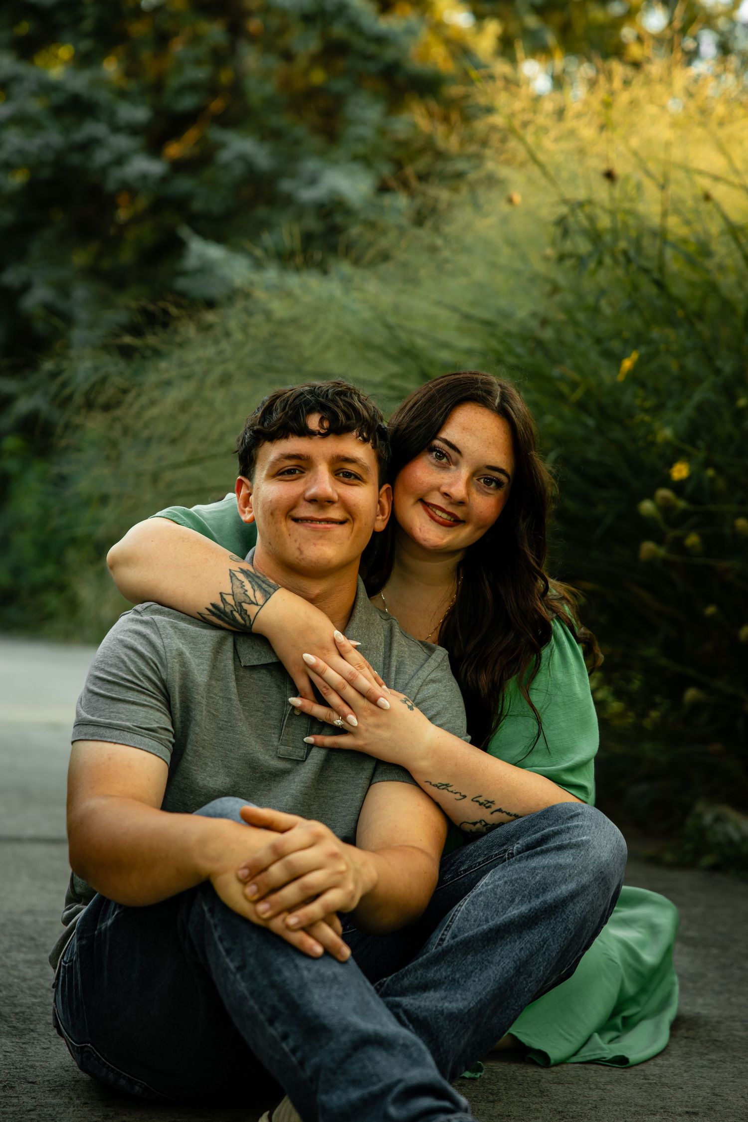 A young couple sitting on pavement, smiling, with the woman hugging the man from behind, surrounded by lush greenery.