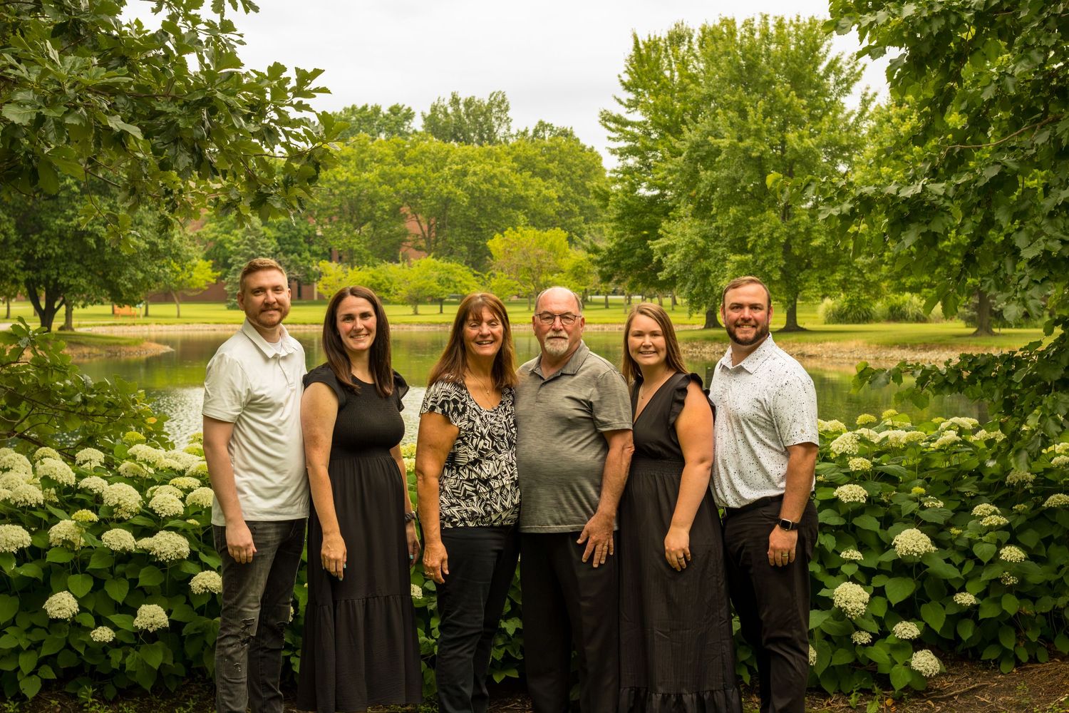 Family of six posing outdoors among white hydrangeas and lush green trees near a serene pond.