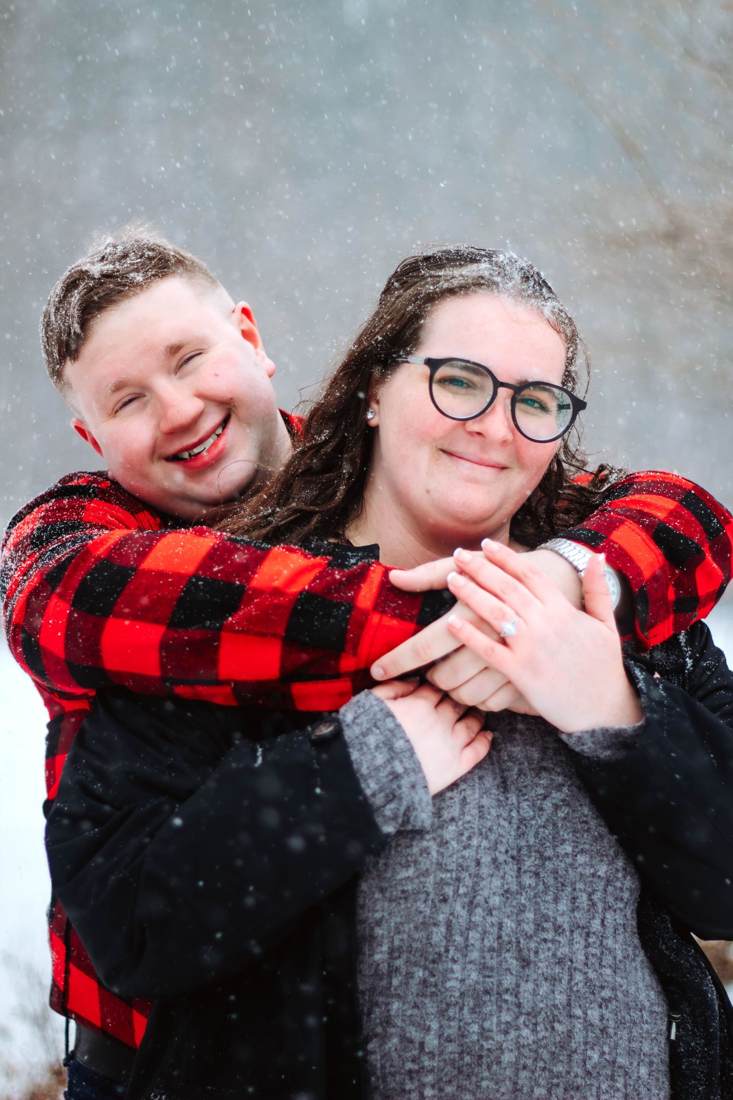 A couple embraces in the falling snow wearing matching buffalo plaid and black winter outfits.