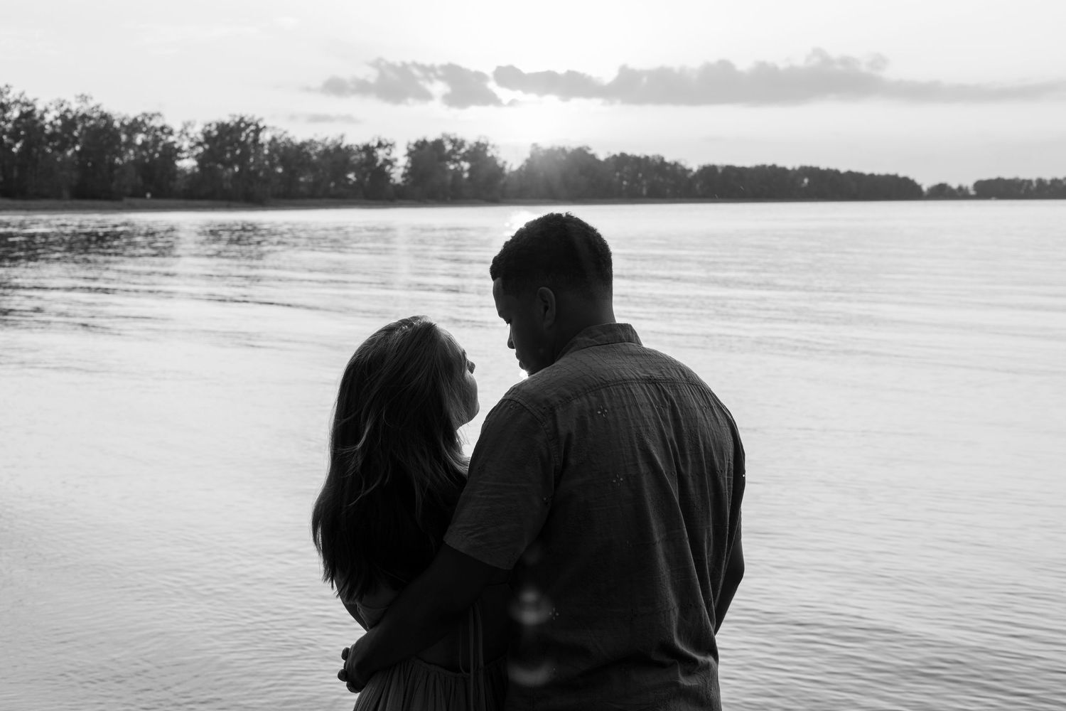 Black and white photo of a couple embracing by a lake at sunset with trees in the background.