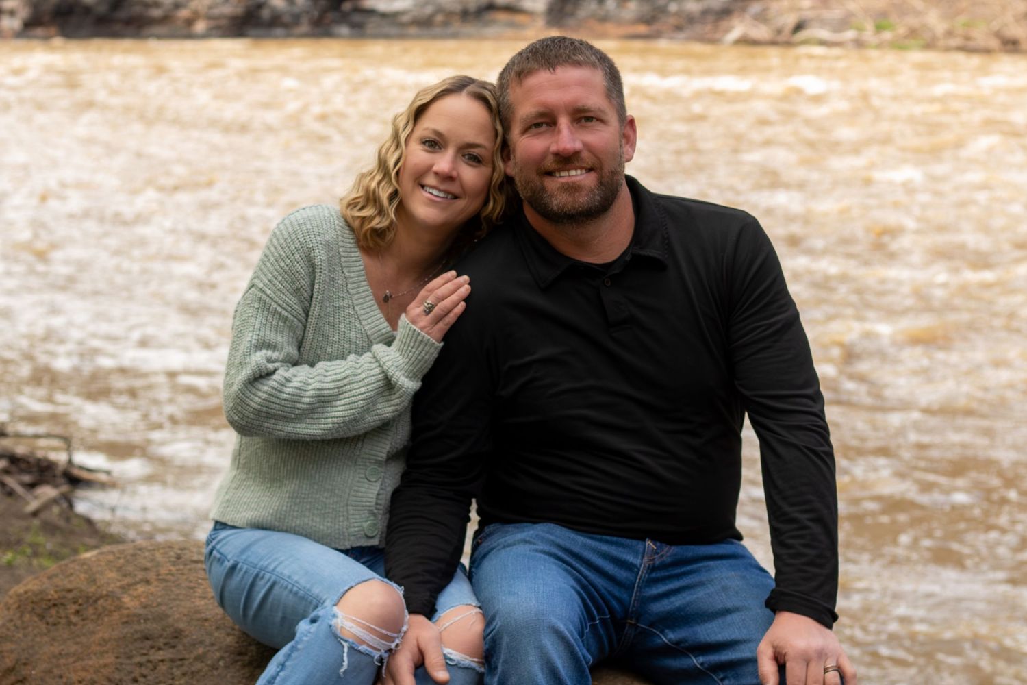 A smiling couple sits together on rocks by a rushing river, woman in green sweater and man in black shirt.