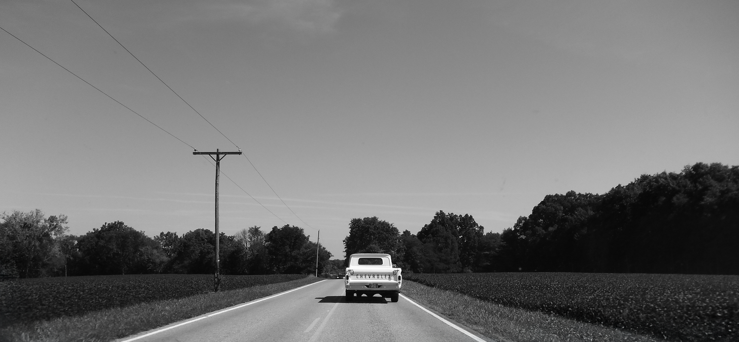 A black and white panoramic view of a rural road stretching to the horizon with a distant car.