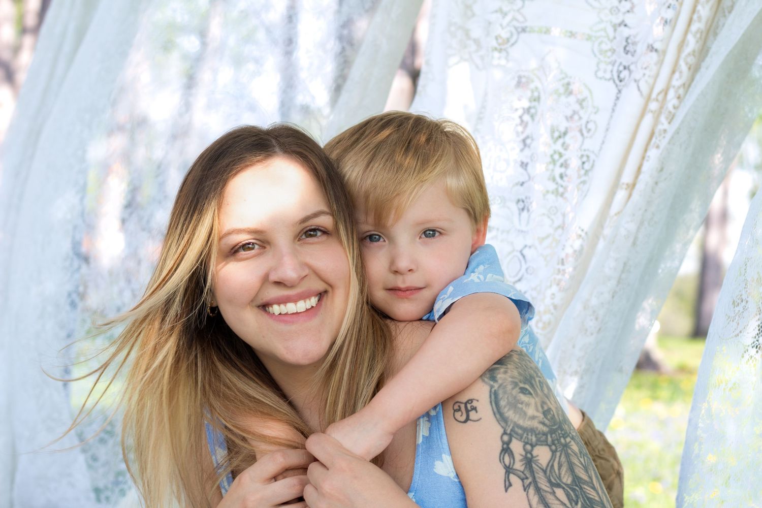 A smiling mother with a tattoo hugs her young blonde child outdoors among white lace curtains in natural light.