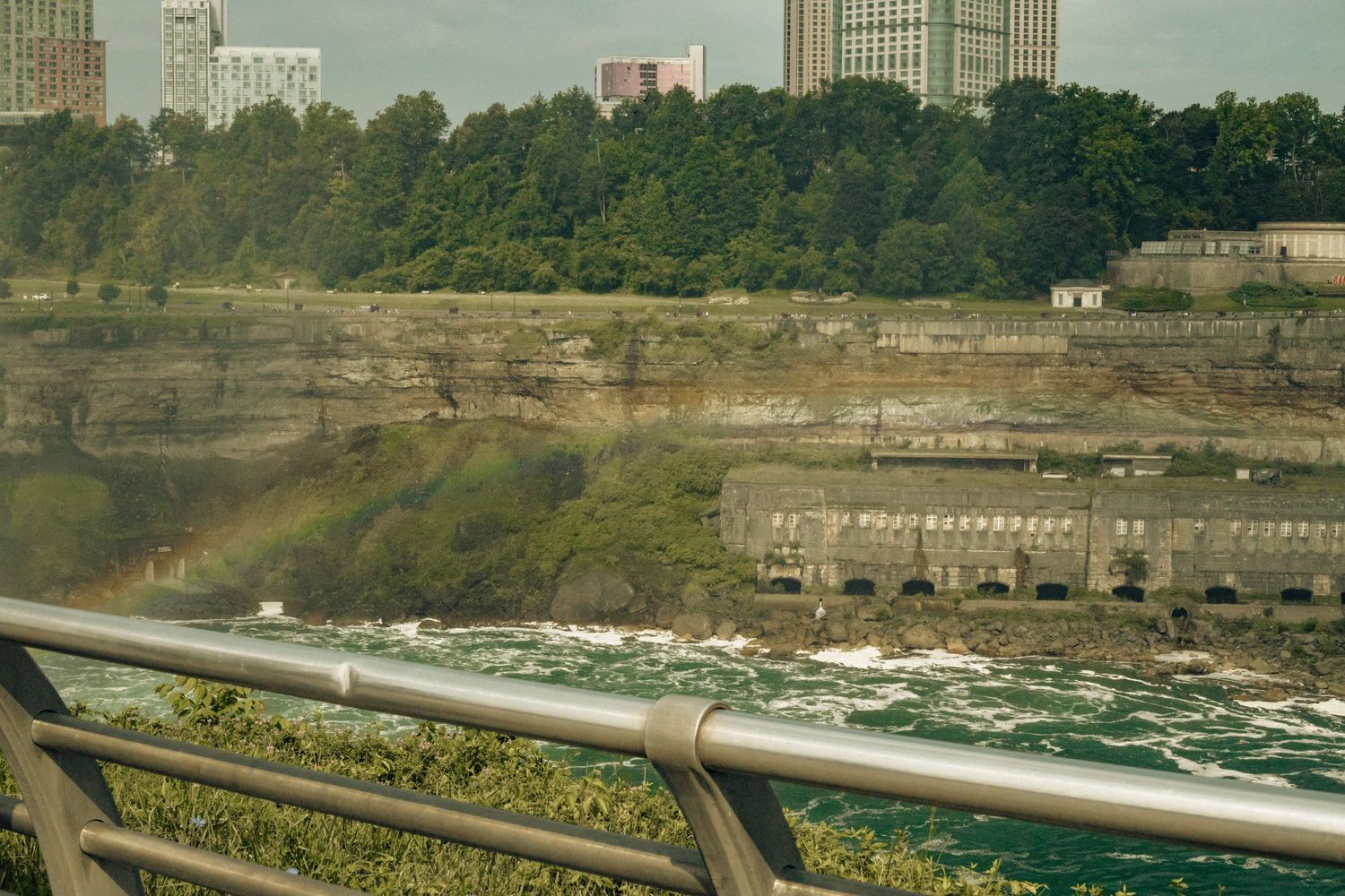 Aerial view of Niagara Falls with a rainbow over rushing rapids, industrial building, lush trees, and city skyline in background.