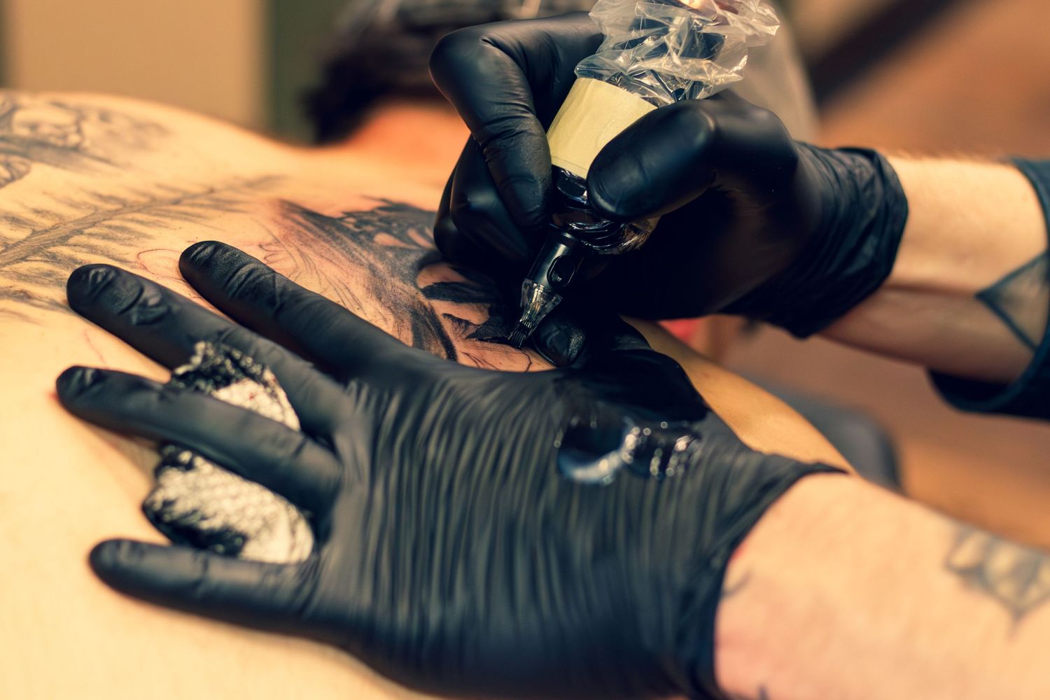 Tattoo artist wearing black gloves carefully inking a detailed tattoo design on a client's skin with a tattoo machine.