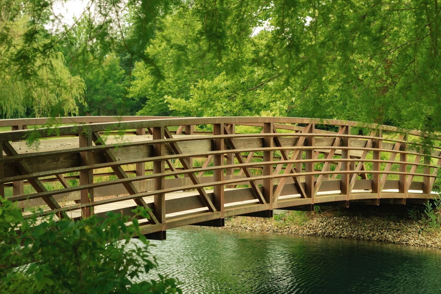 A wooden arched footbridge spans a calm green river surrounded by lush trees in a peaceful park setting.