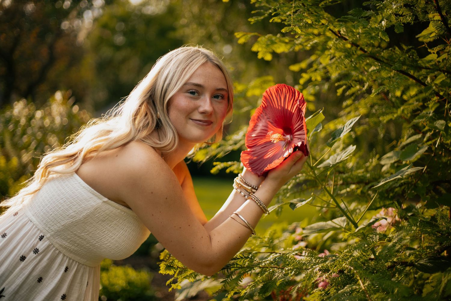 Smiling blonde woman in white dress holding a red flower among lush green bushes in a garden setting.