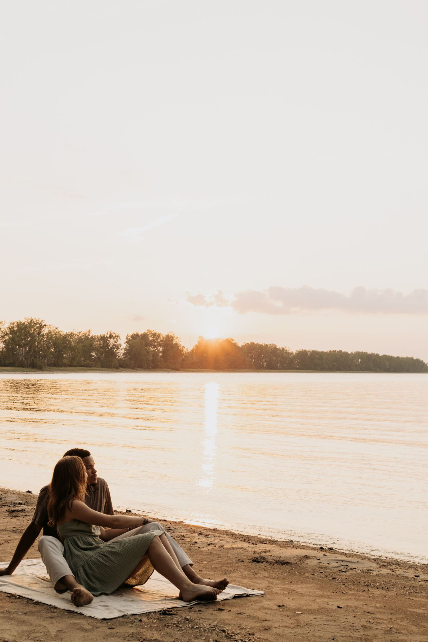 Couple sitting on sandy beach blanket watching golden sunset reflect over calm river with trees silhouetted in background.