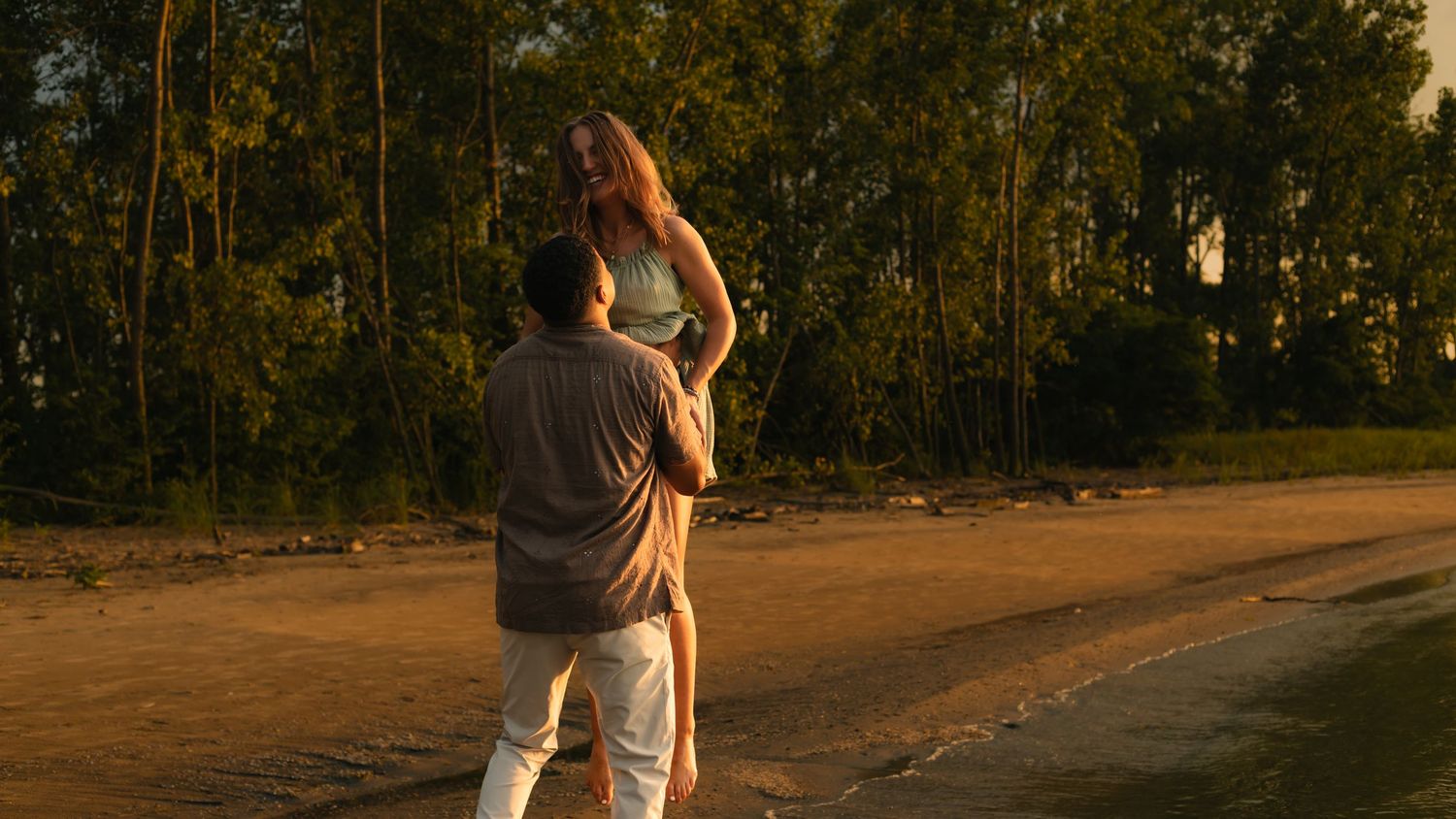 Couple sharing a romantic moment on a sandy beach at golden sunset with lush green trees in background.