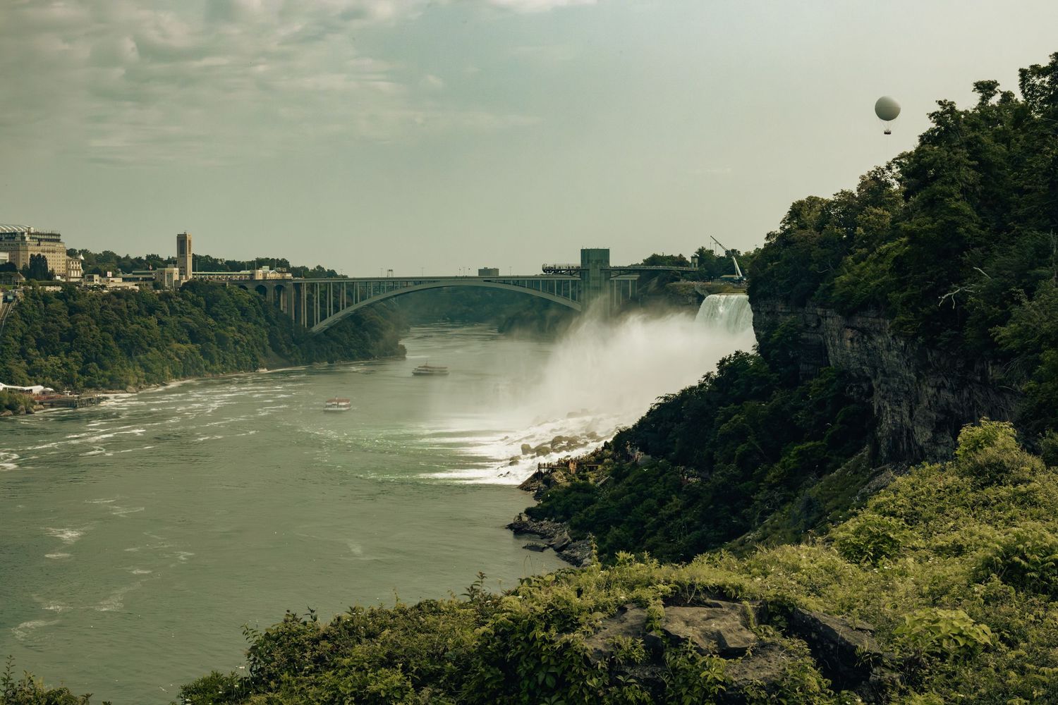 Aerial view of Niagara Falls with mist rising, lush greenery, and a bridge spanning the river under a cloudy sky.