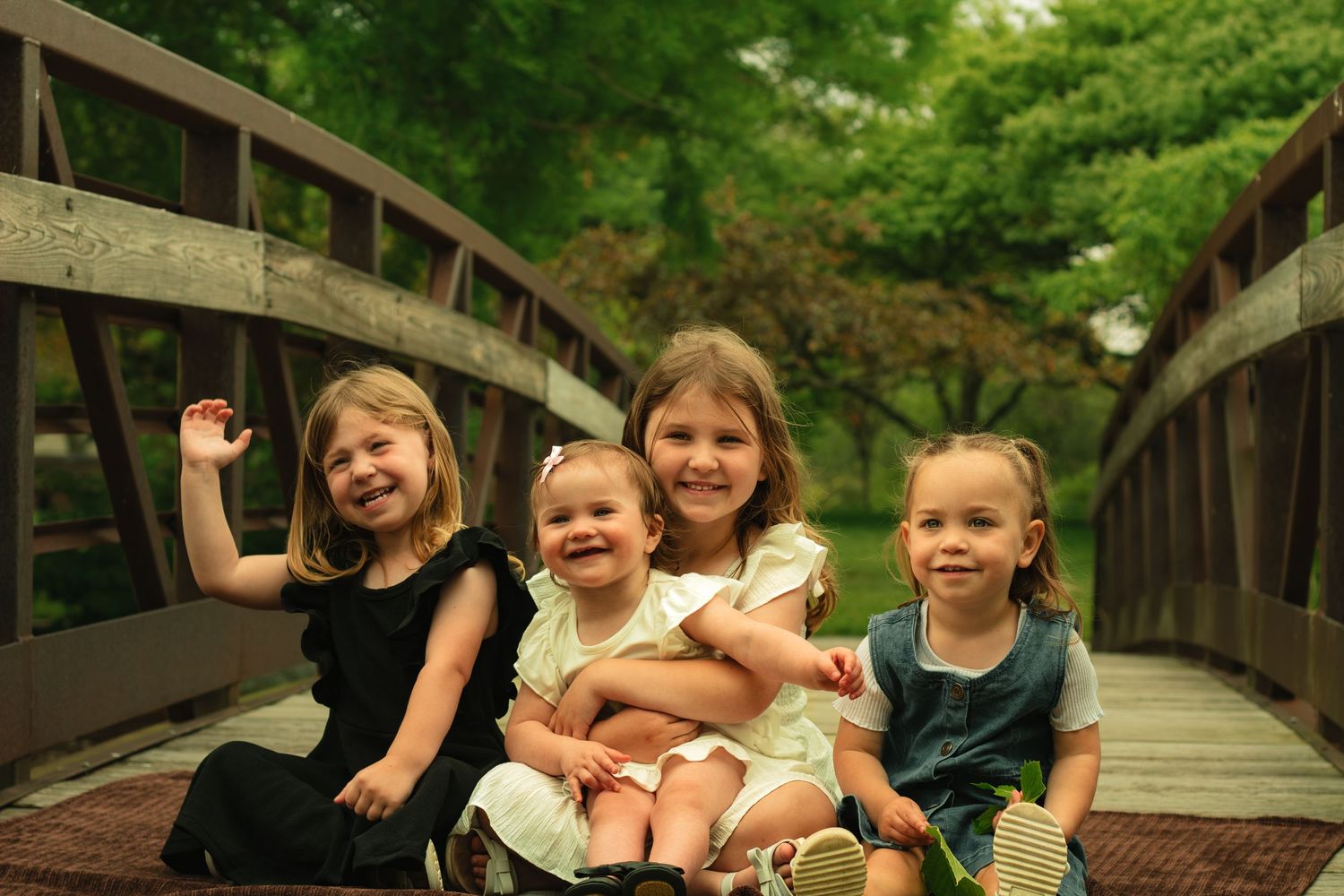 Four young girls smiling and posing together on a wooden bridge surrounded by lush green trees.