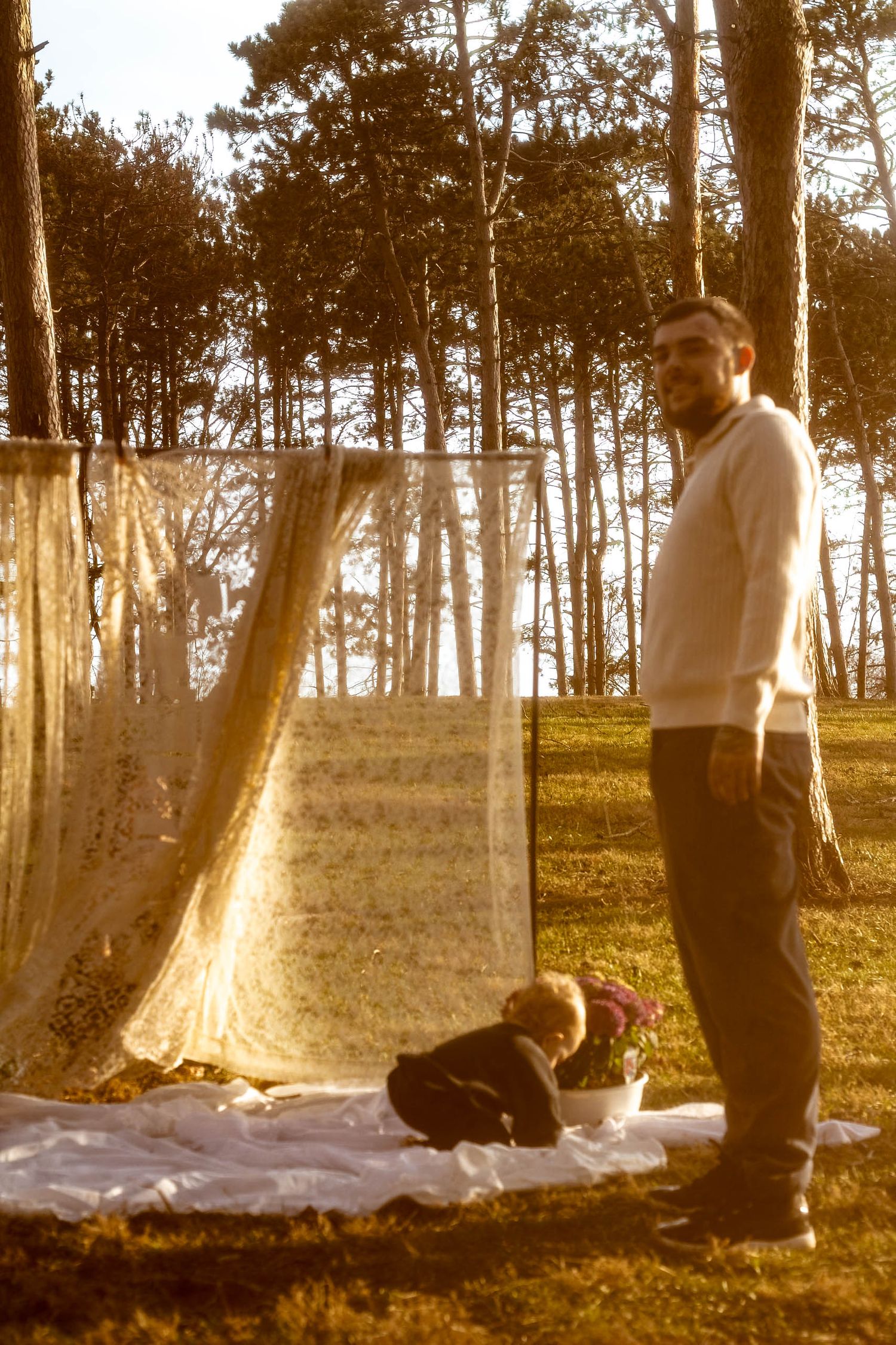 A vintage sepia photograph shows someone standing next to a blanket laid out on grass with a small dog in a wooded setting.