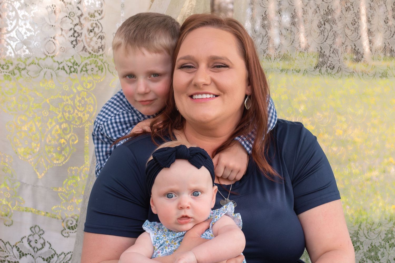 A smiling woman holds a baby girl while a young boy stands behind her outdoors.