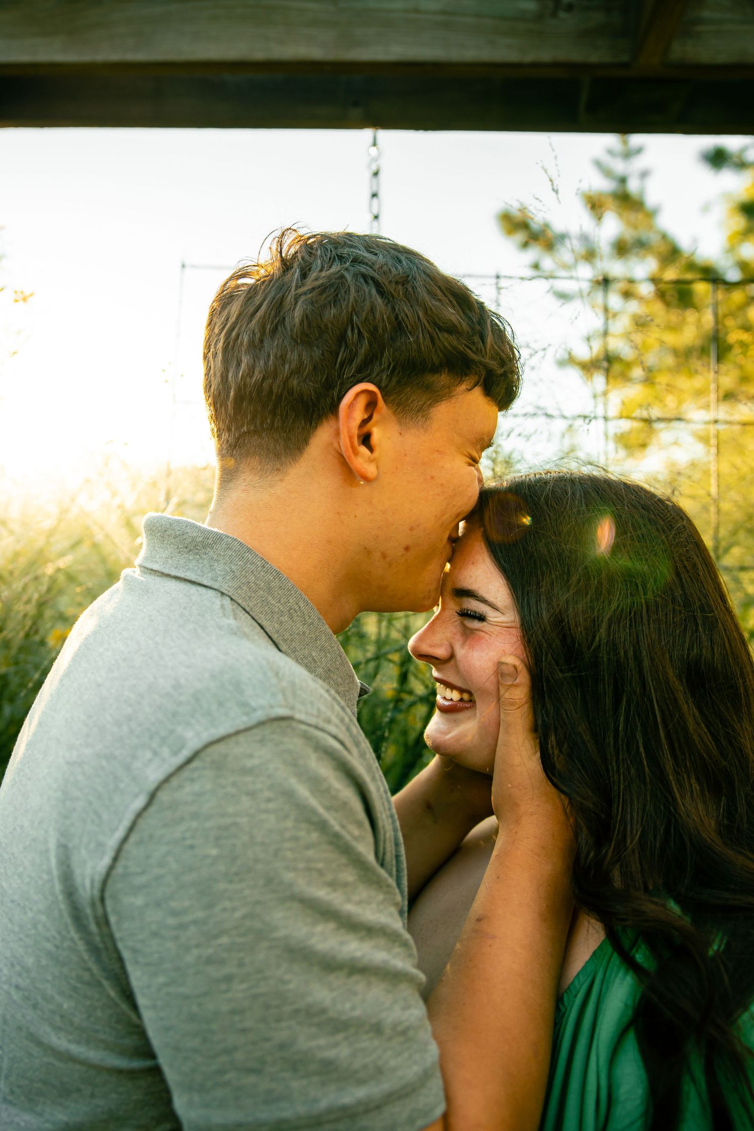 A young couple sharing a tender moment outdoors, laughing together in golden sunlight near lush greenery.