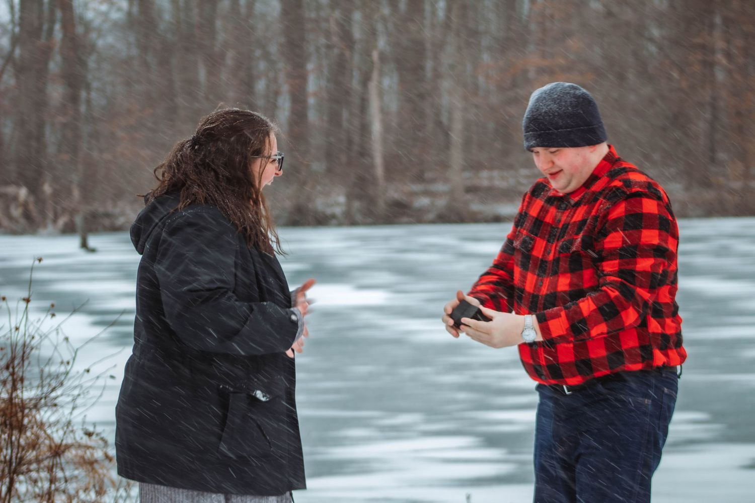 A couple stands by a frozen lake in winter, with one person wearing a red buffalo plaid jacket and black beanie.
