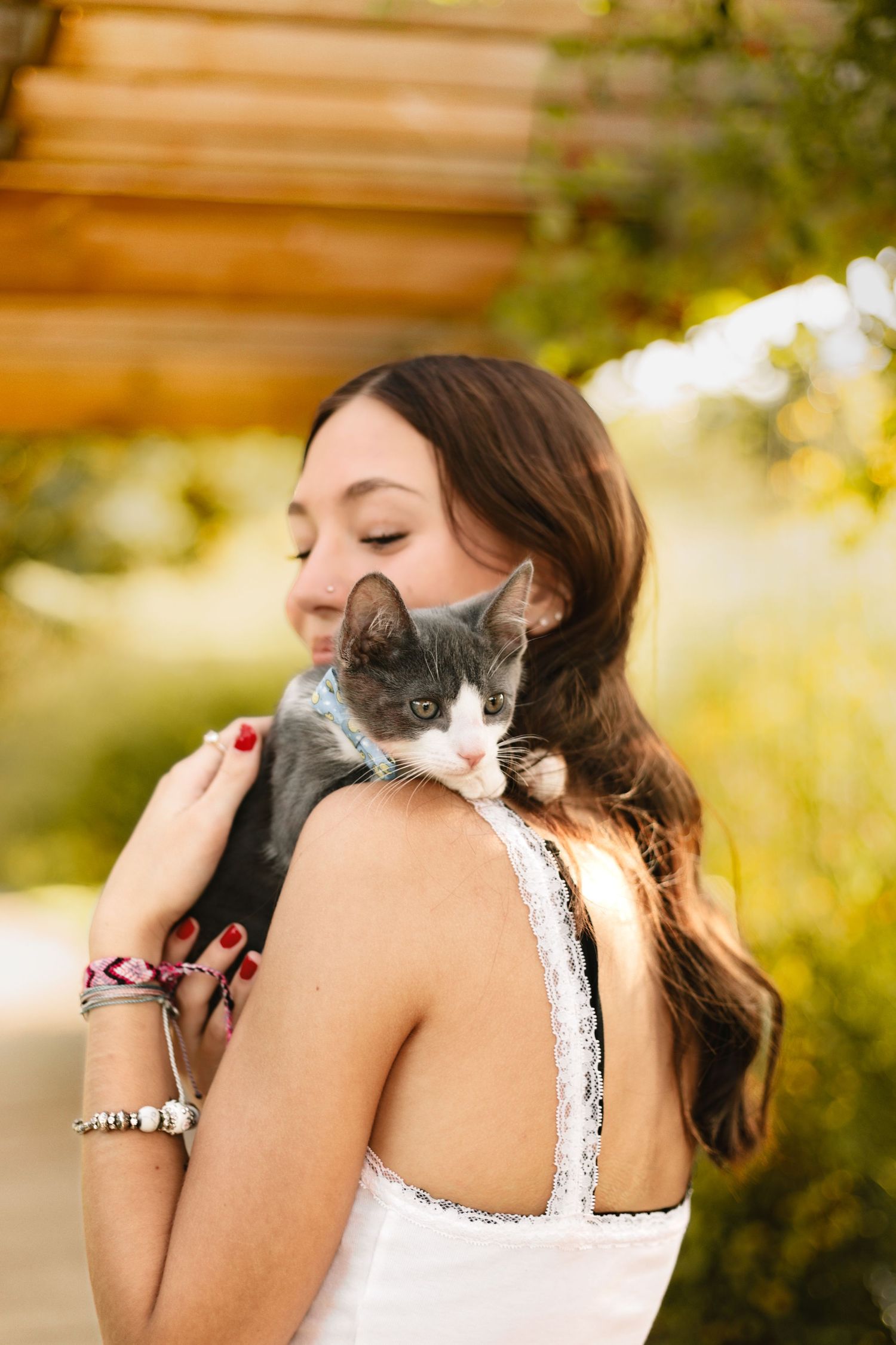 Young woman with long brown hair tenderly holds a gray and white kitten on her shoulder outdoors in warm sunlight.