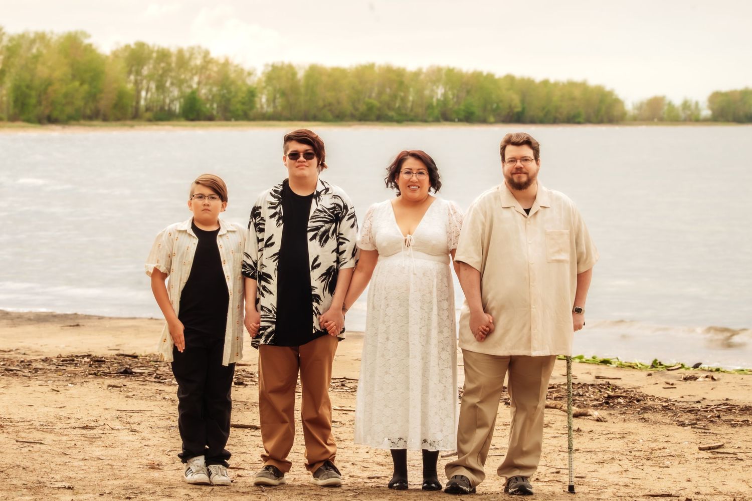 Family of four posing on a sandy riverbank with lush green trees and calm water in the background.