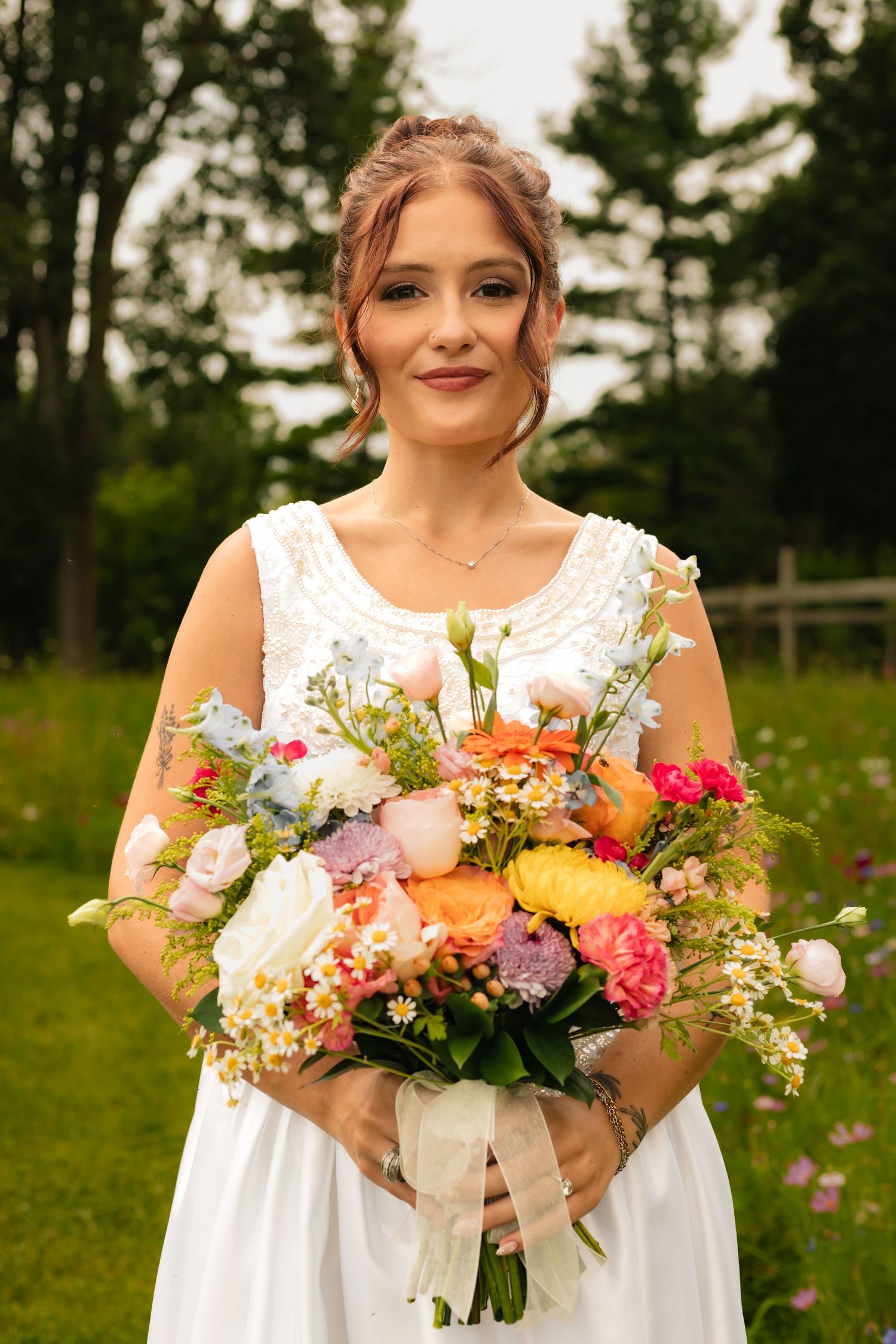 Bride in white dress holding a colorful wildflower bouquet outdoors with green trees in the background.