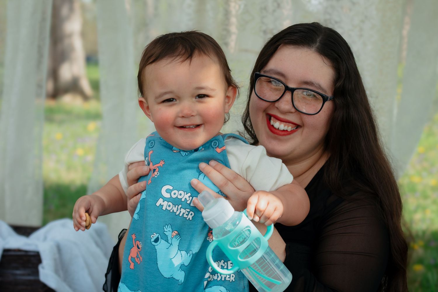Smiling woman with glasses holding a happy baby wearing a Cookie Monster bib outdoors.