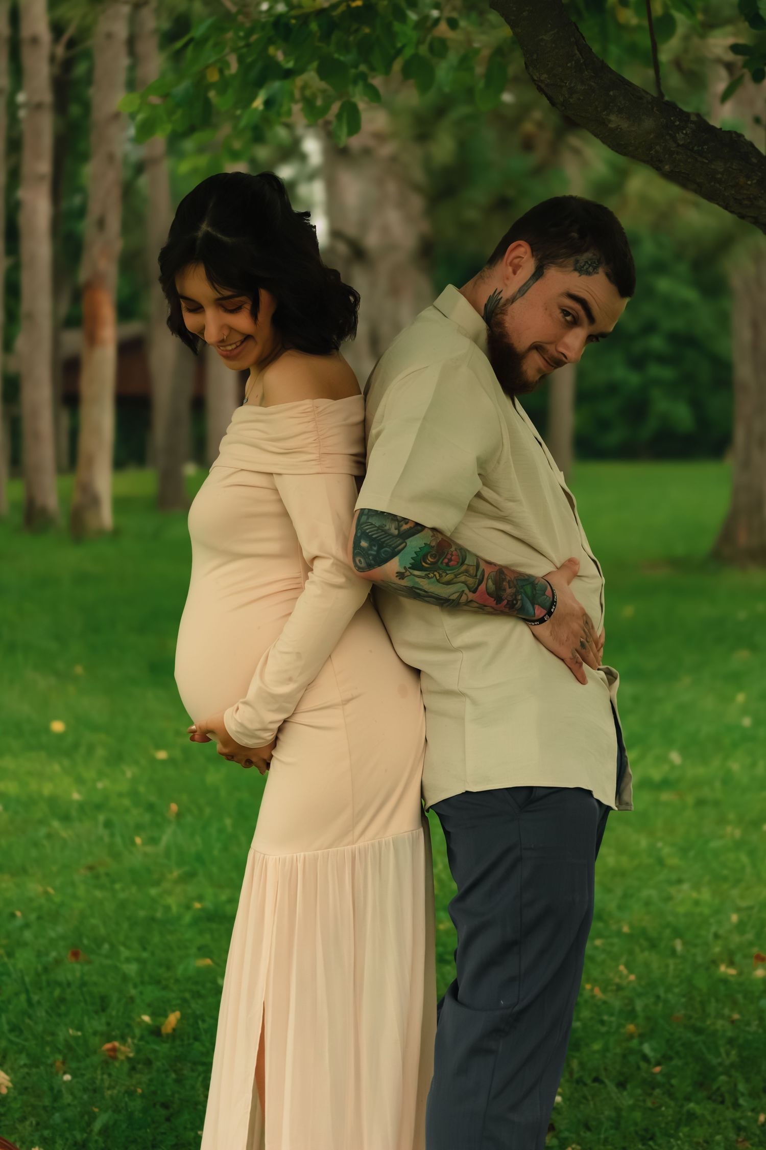 Pregnant woman in pink dress and tattooed man stand back-to-back in a lush green park during a maternity photoshoot.