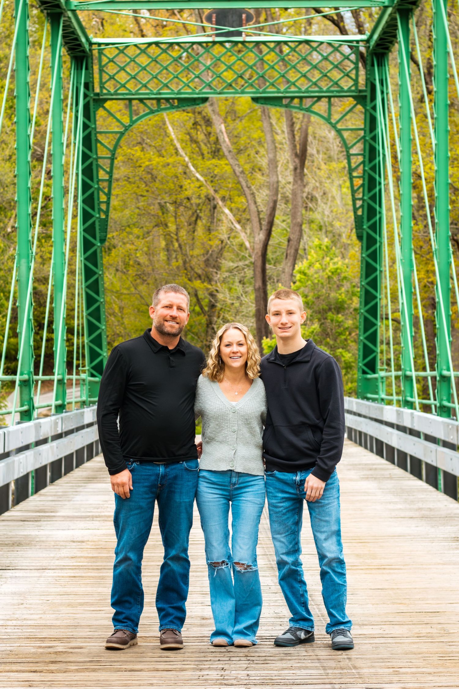 Family of three posing on a green metal bridge surrounded by lush trees in a warm outdoor portrait.