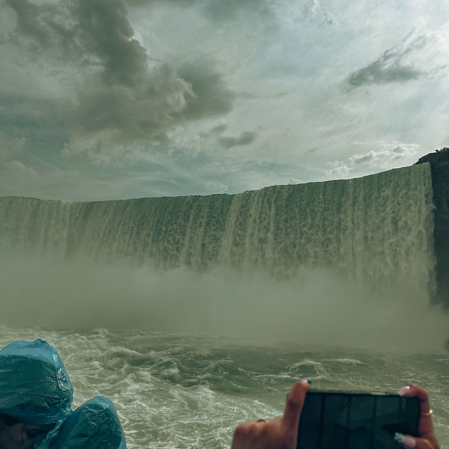 Tourists in blue rain ponchos photographing massive Niagara Falls waterfall from a boat amid misty spray under cloudy skies.