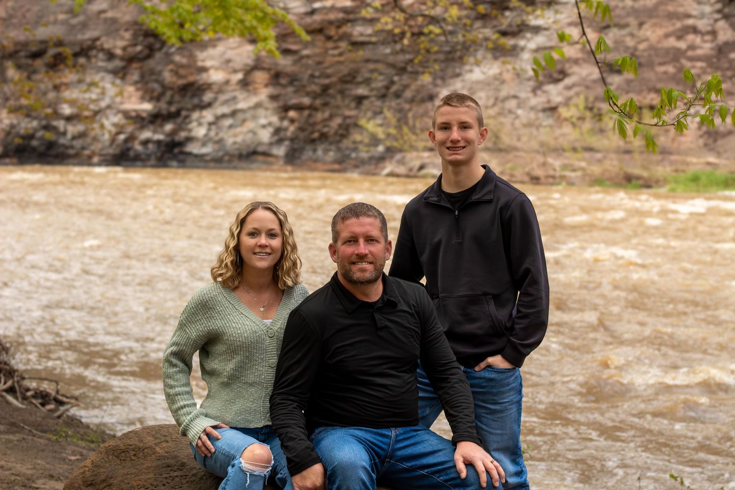 Family of three posing by a rushing river with rocky cliffs, smiling outdoors in casual attire.