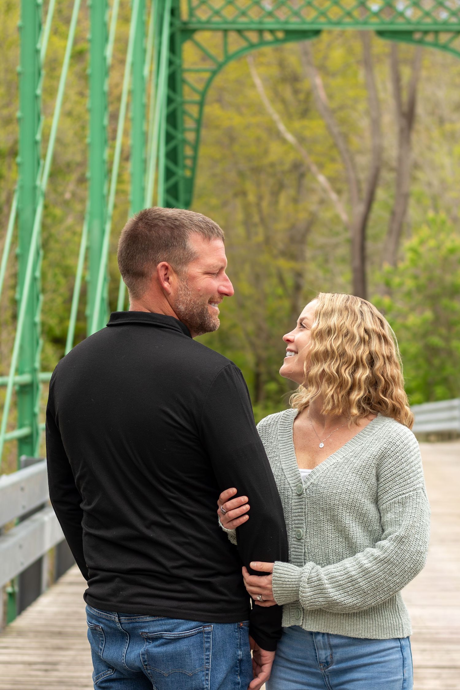 A smiling couple shares a tender moment on a green iron bridge surrounded by lush trees in spring.