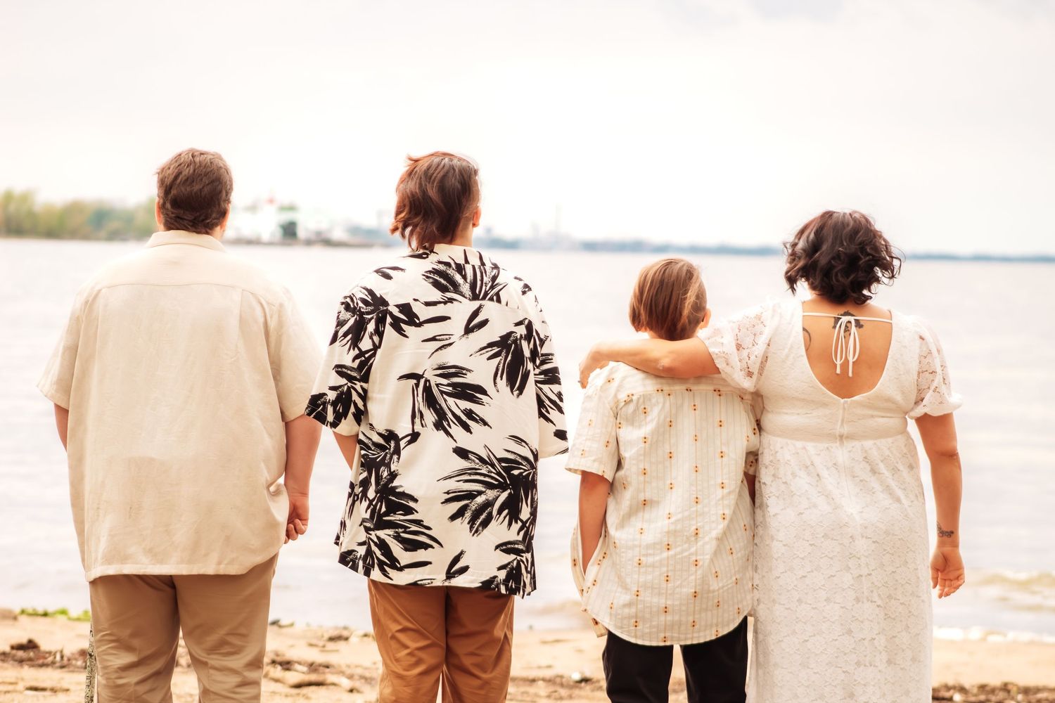 Four people standing together facing a calm lake shore, viewed from behind on a sandy beach.