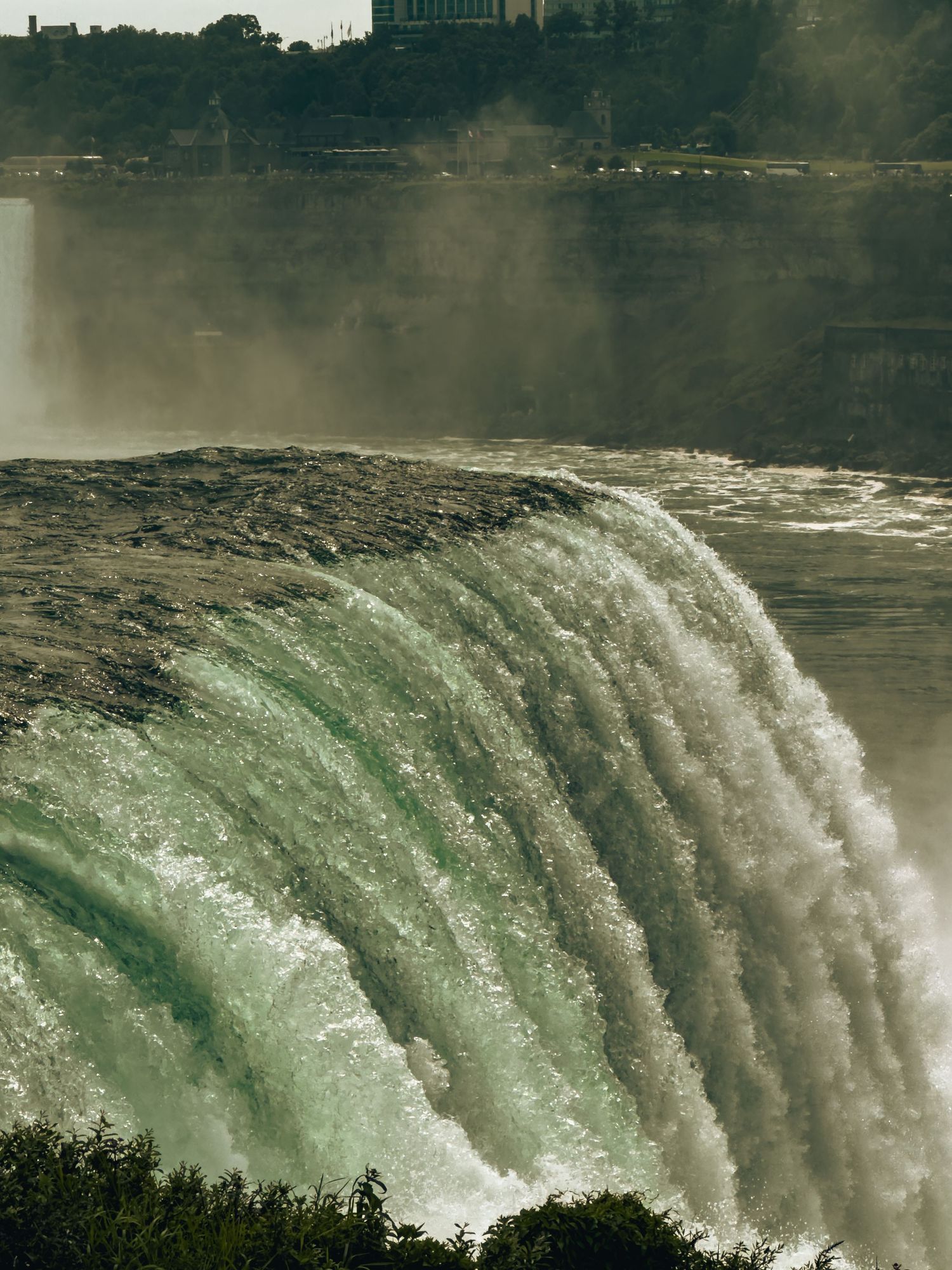Aerial view of Niagara Falls Horseshoe waterfall with green rushing water and mist rising against a hazy skyline.