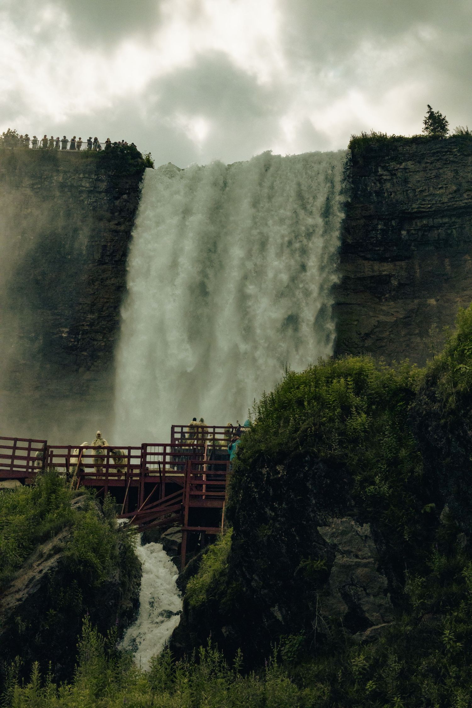 Massive waterfall cascades down rocky cliffs with a wooden viewing platform and lush greenery under dramatic cloudy skies.