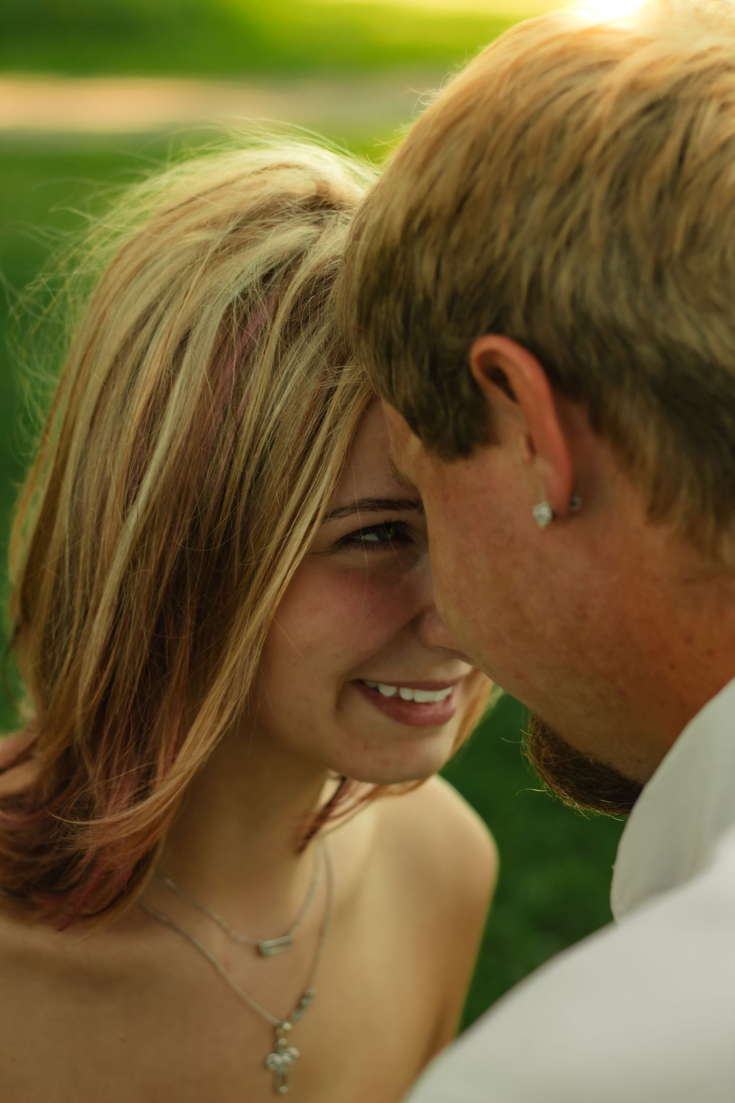 A smiling young woman with highlighted hair wearing a necklace leans close to a man outdoors in soft sunlight.