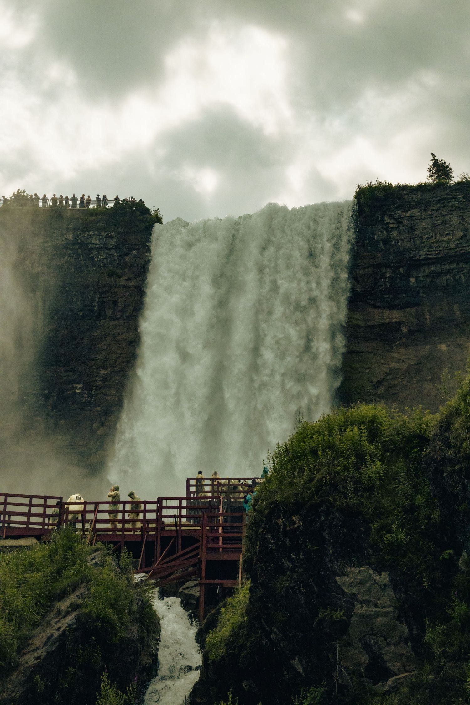Tourists on a wooden viewing platform observe a massive waterfall cascading down rocky cliffs under overcast skies.