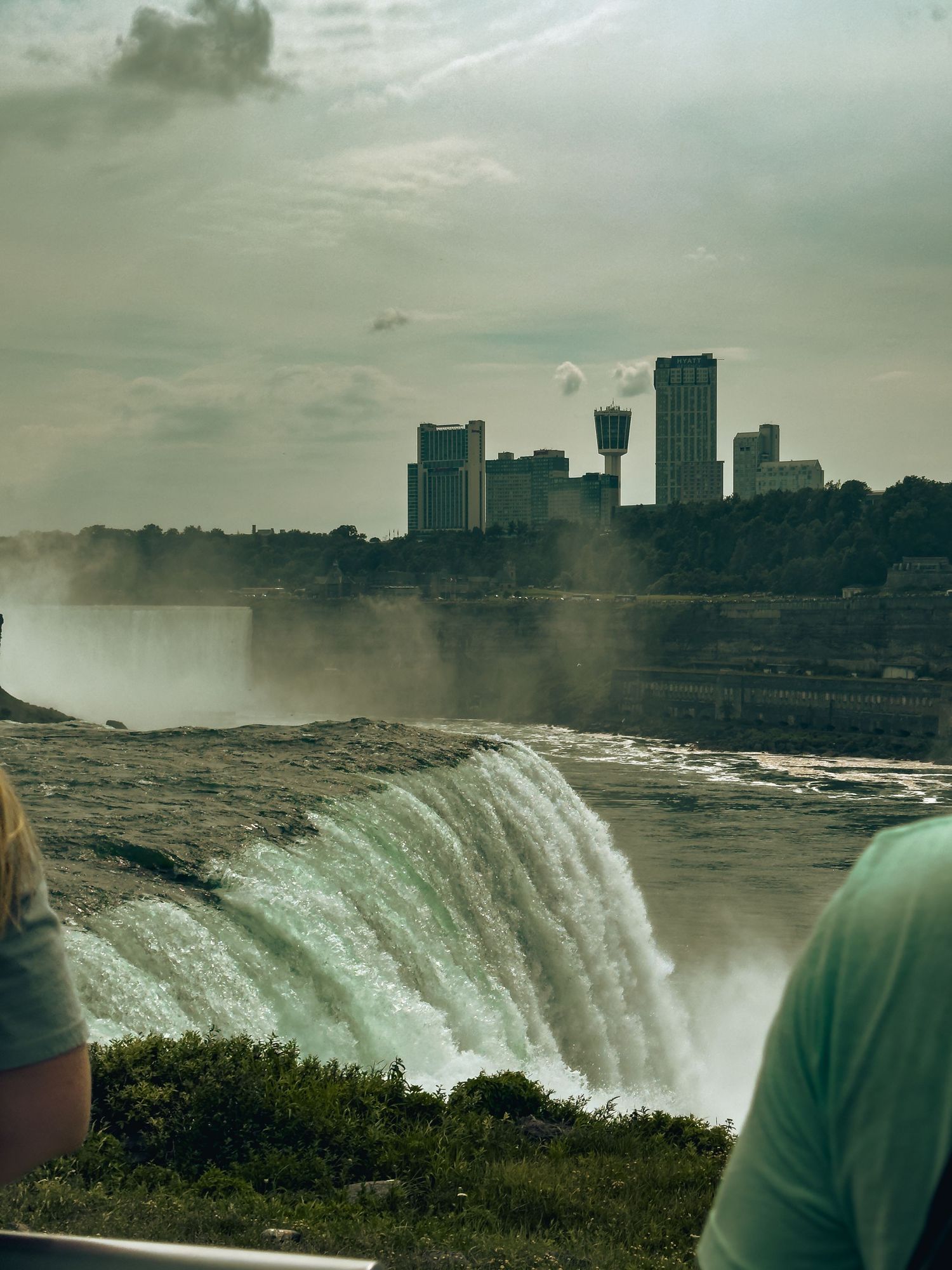 Tourists viewing Niagara Falls with powerful cascading water and Niagara city skyline visible in the misty background.
