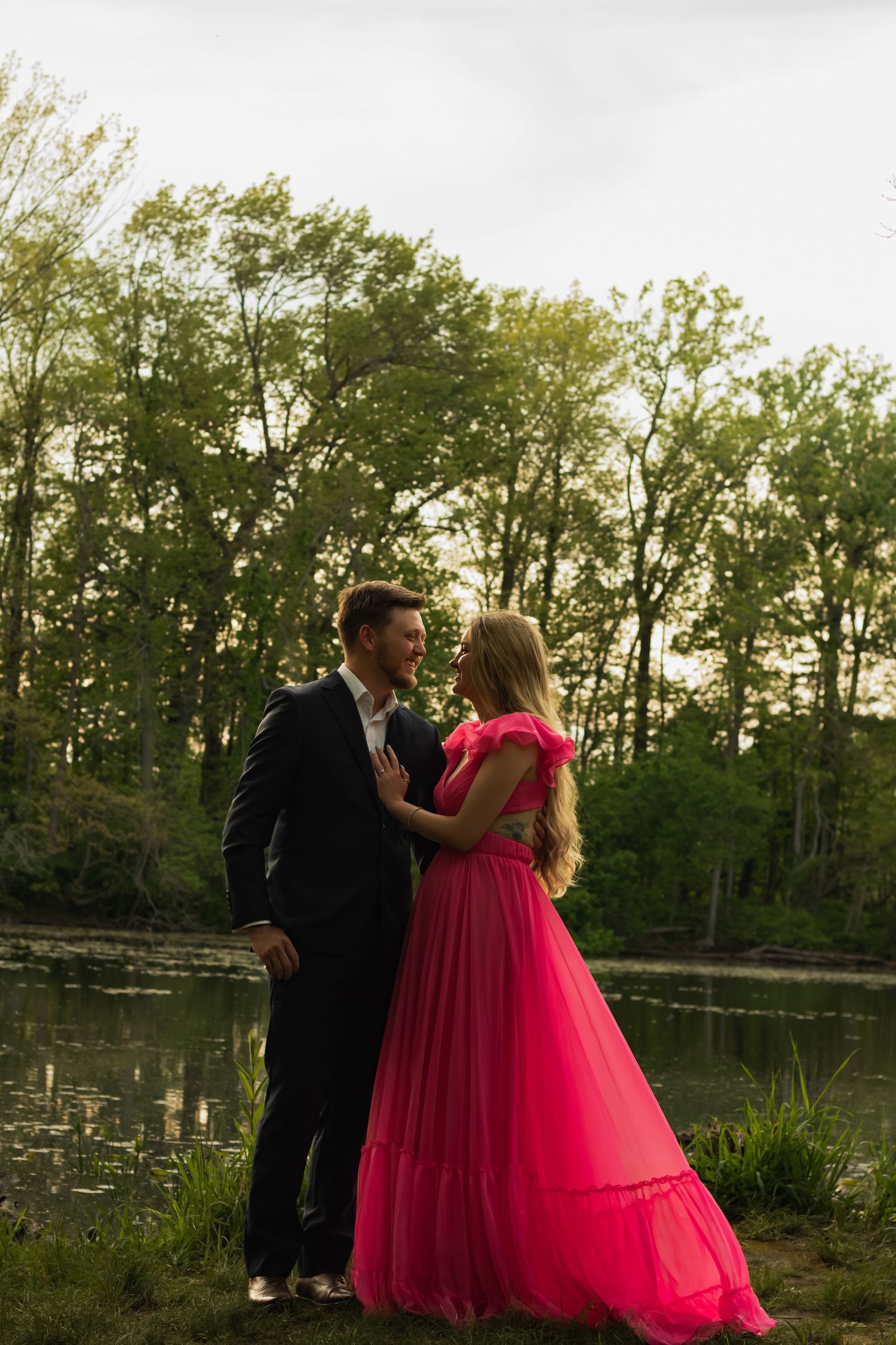 Couple in formal wear poses by a pond, woman in pink gown, man in dark suit, surrounded by lush green trees.