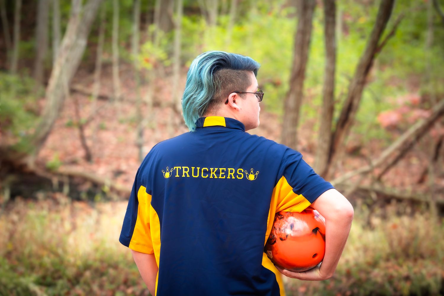 Person wearing a Truckers uniform holds an orange pumpkin in a wooded setting.