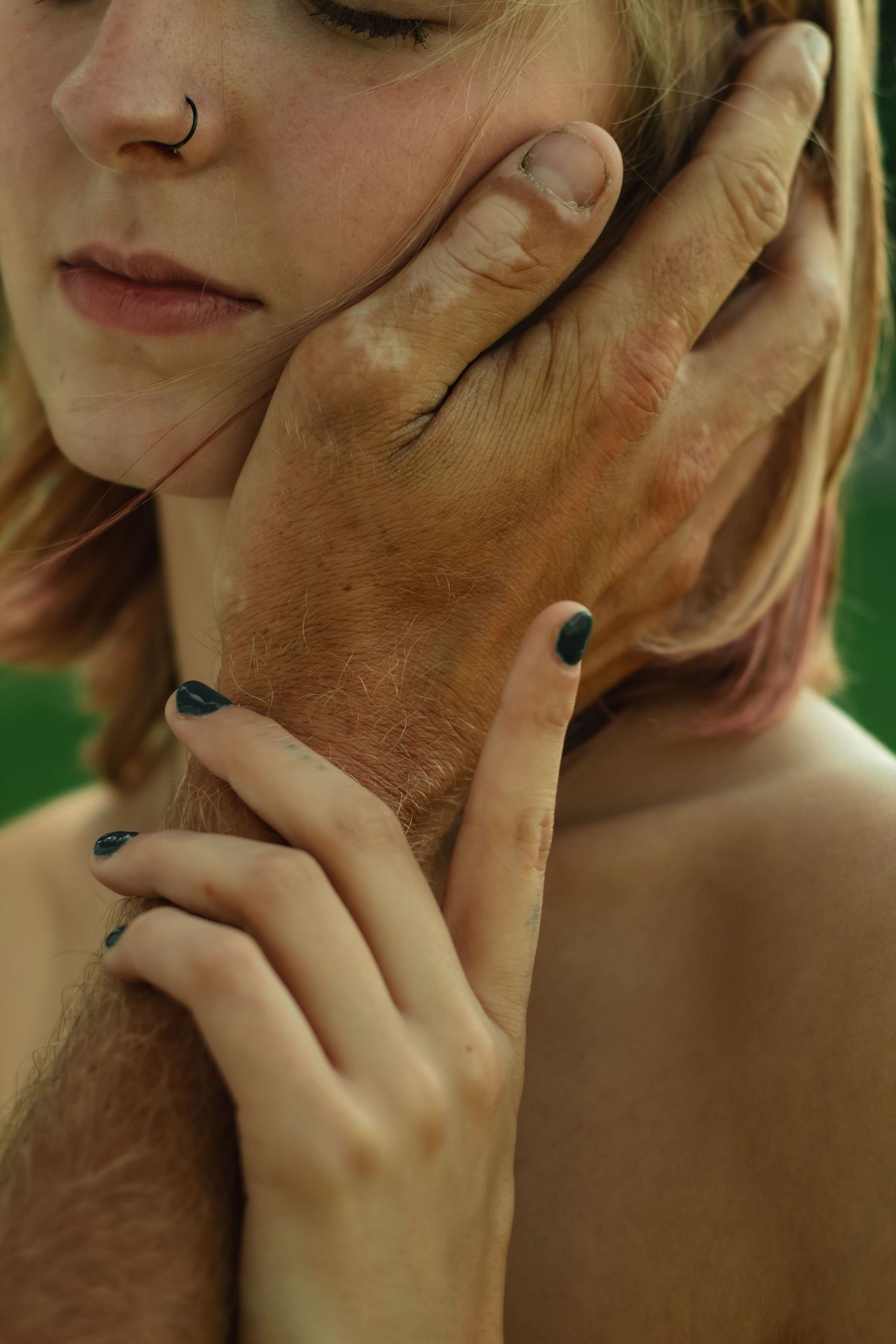 Close-up of a man's weathered hands gently cradling a woman's face, her dark green nails resting on his arm.