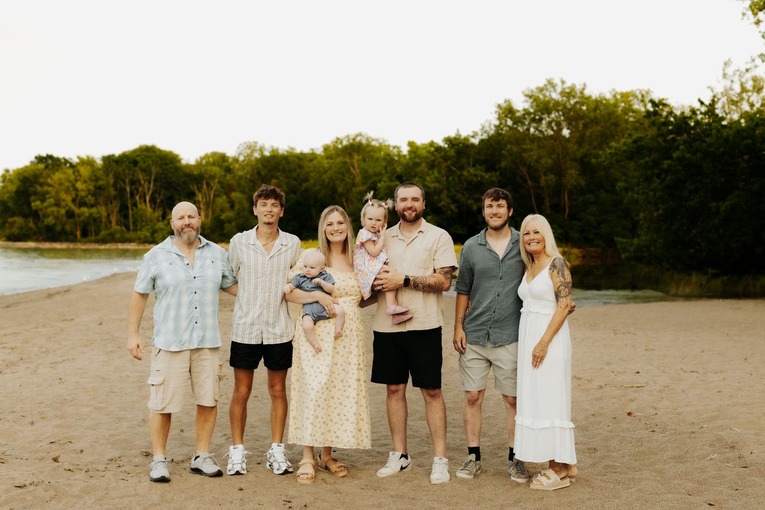 Family of seven posing together on a sandy beach at sunset with lush green trees in the background.