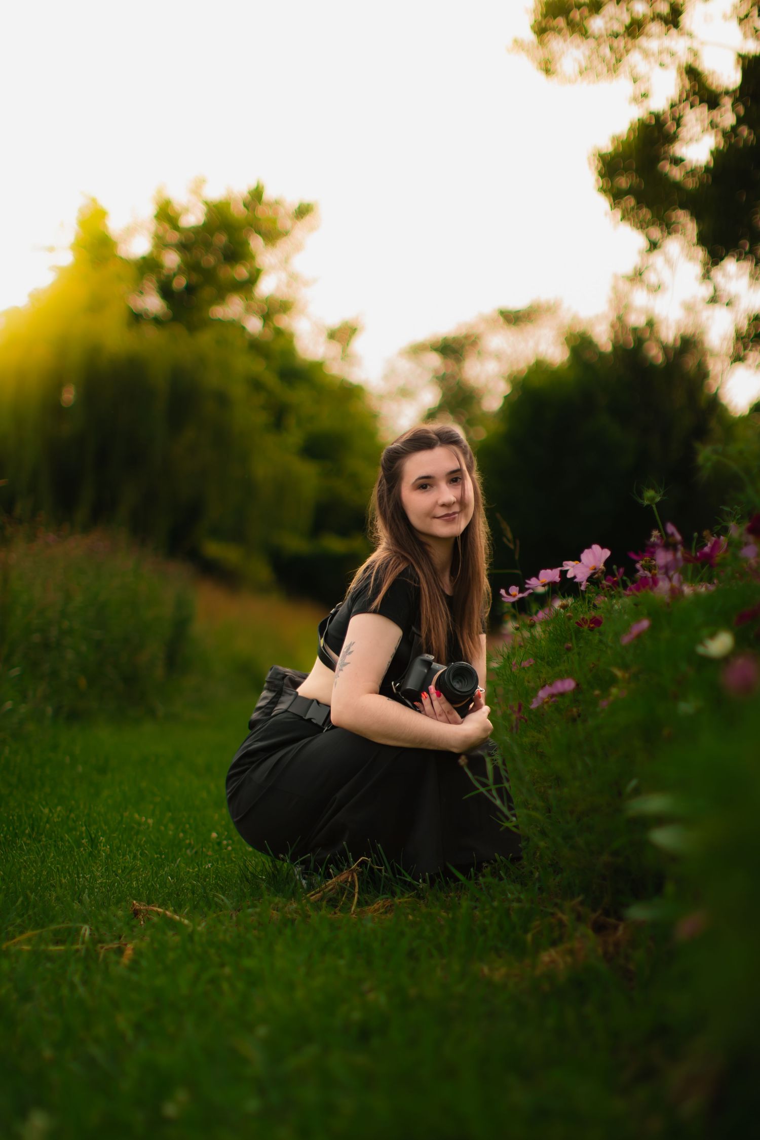 Young woman crouching in a lush green park holding a camera near pink flowers during golden hour sunset.