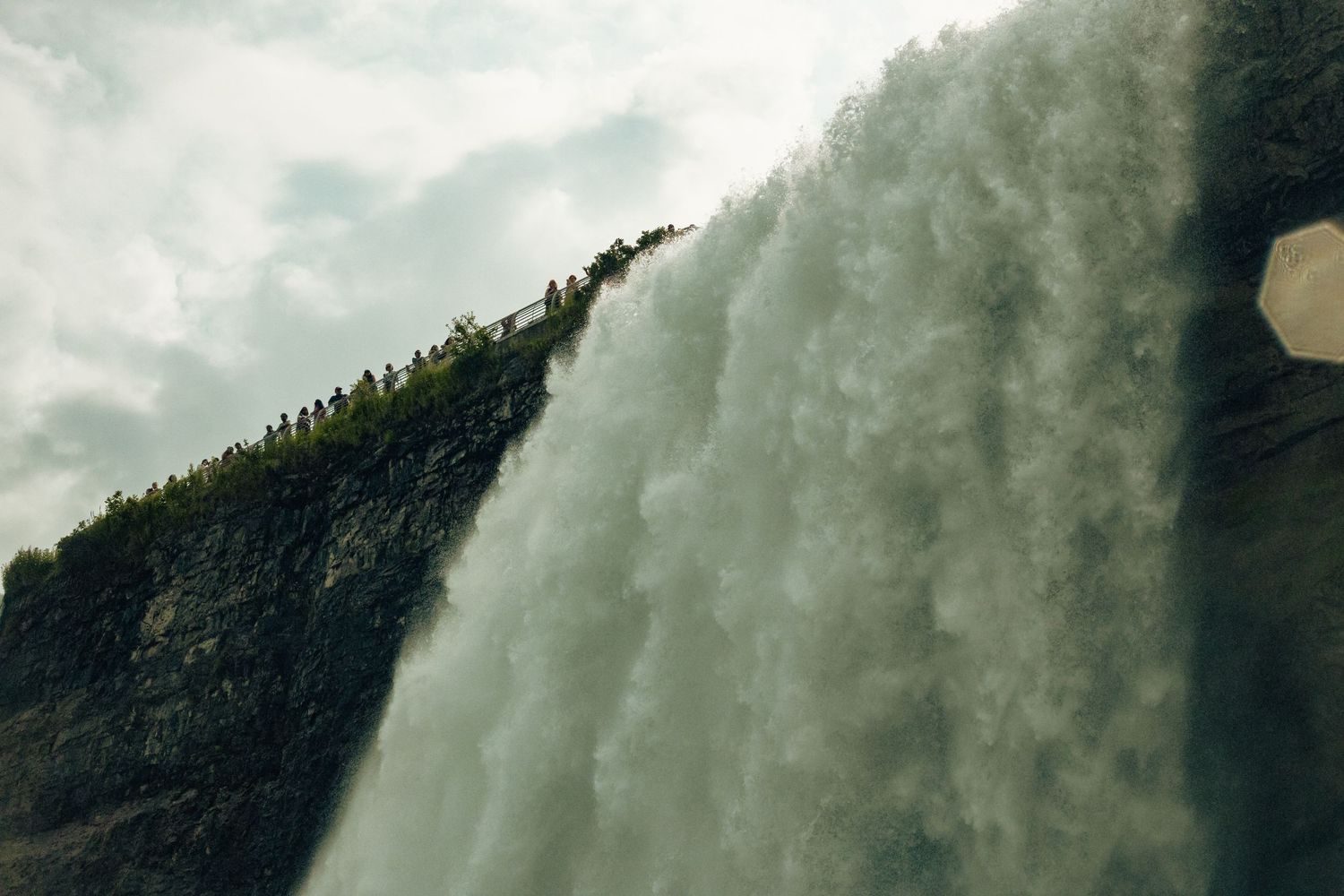 A massive waterfall cascades down a rocky cliff with people visible on the ridge above against a cloudy sky.