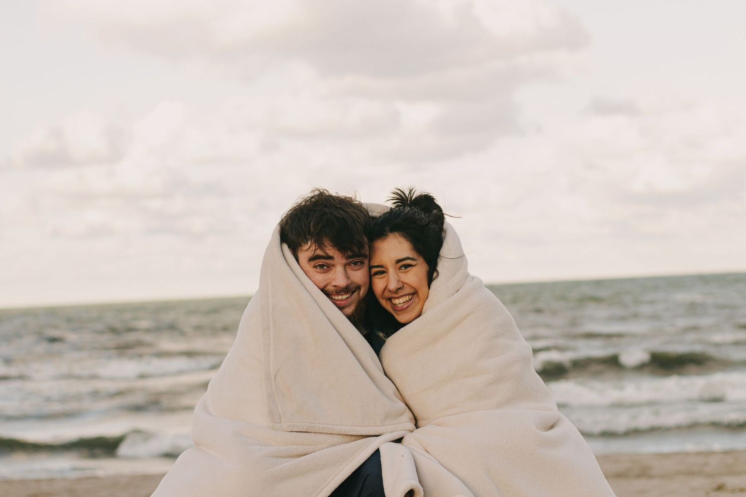 A couple shares intimate moments wrapped in a blanket on a cloudy beach.