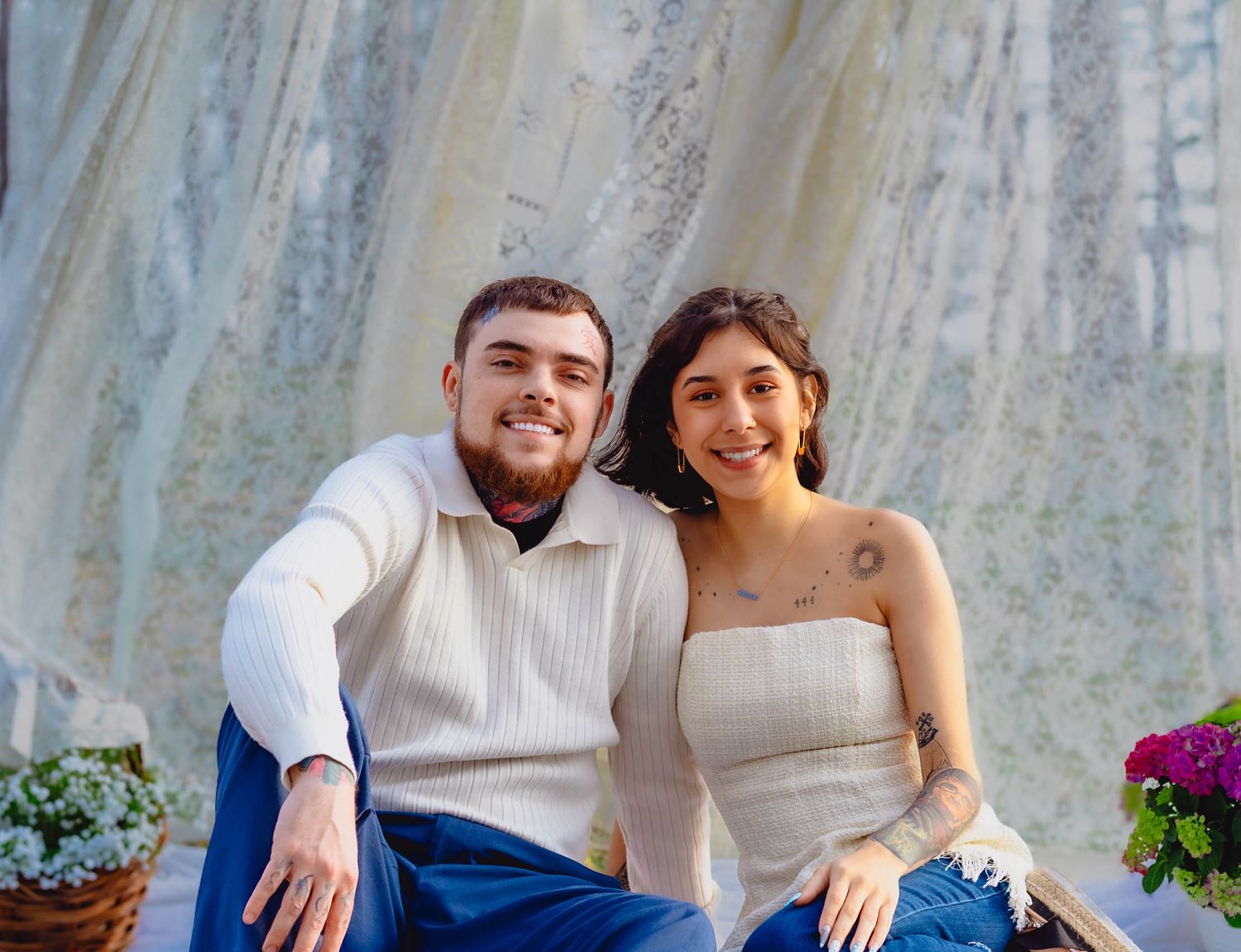 A couple sits together outdoors against a light curtained backdrop with purple flowers visible to the side.