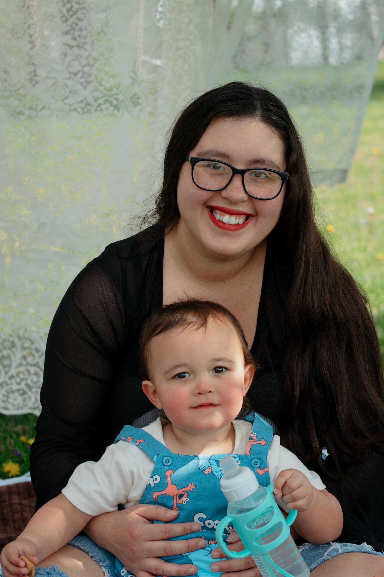 Young woman with dark hair and red lips holds a smiling baby in a blue bib outdoors.