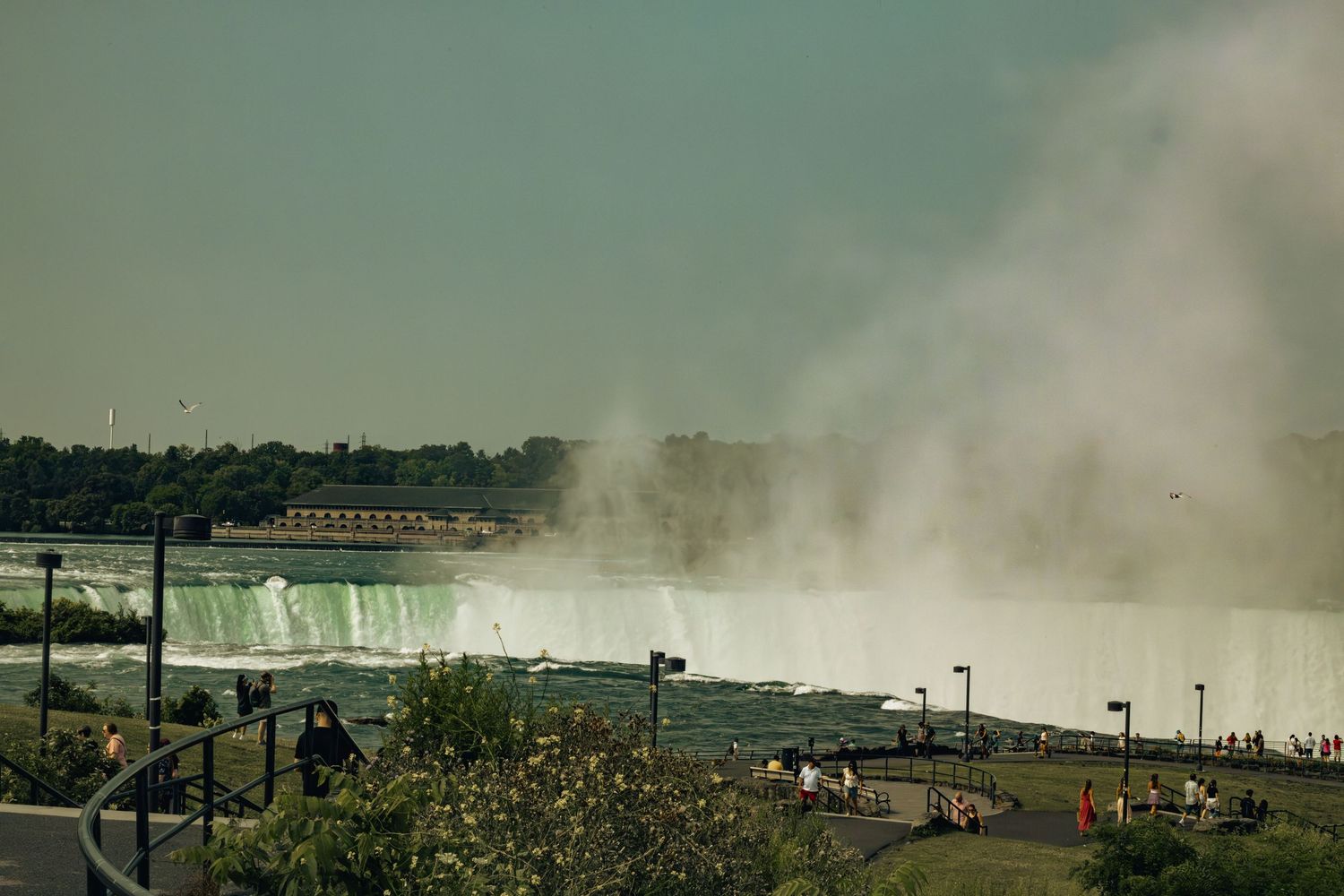 Tourists viewing the misty Niagara Falls from a scenic walkway with lush greenery and a building in the background.